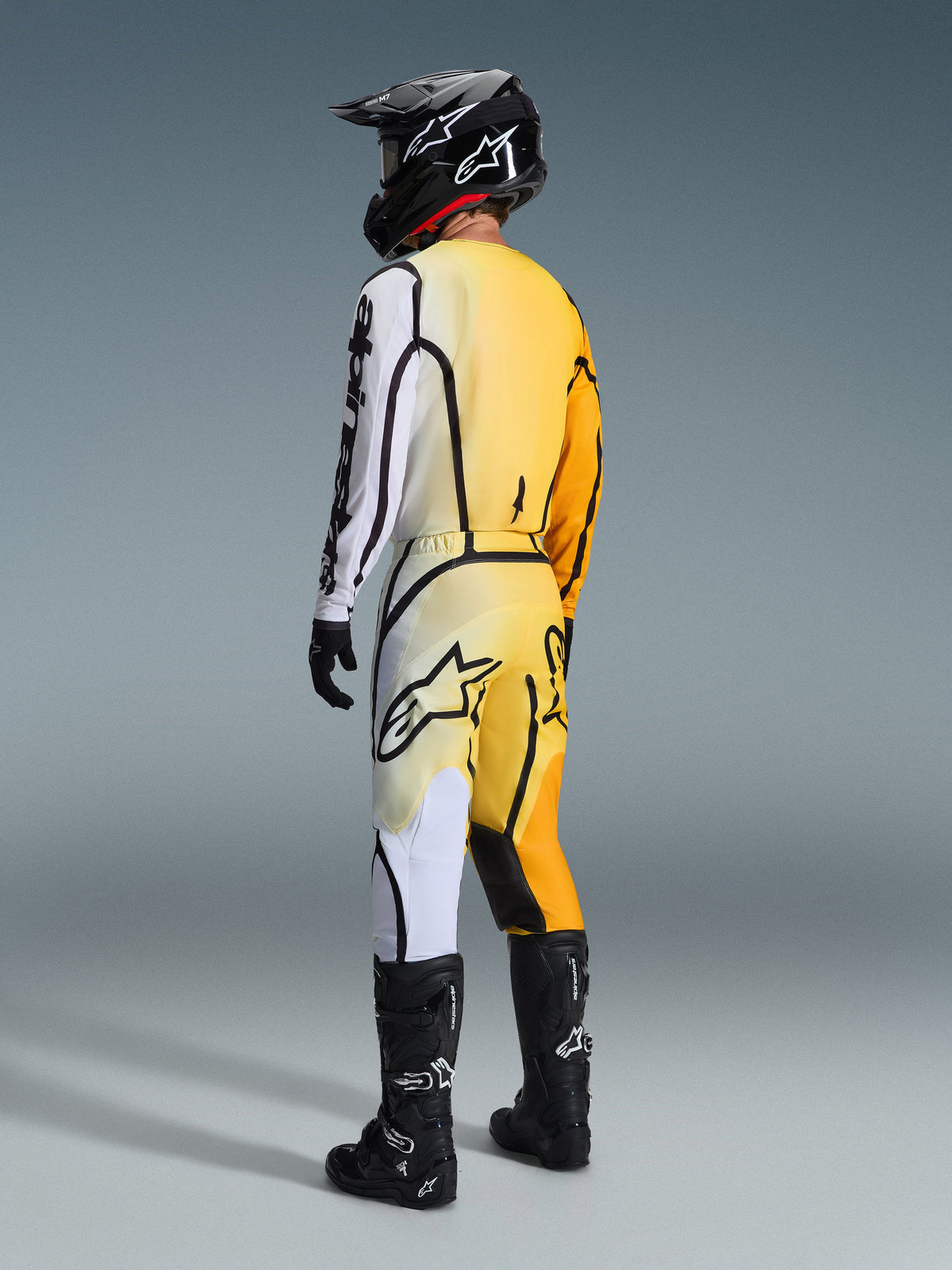 Alpinestars 2026 Fluid Apex Jersey en bijbehorende broek, motorcrosser van achteren gezien in een geel-zwarte outfit met geel-naar-wit kleurverloop en opvallende zwarte grafische lijnen, gecombineerd met een zwarte helm, zwart-witte handschoenen en zwarte motorcrosslaarzen.