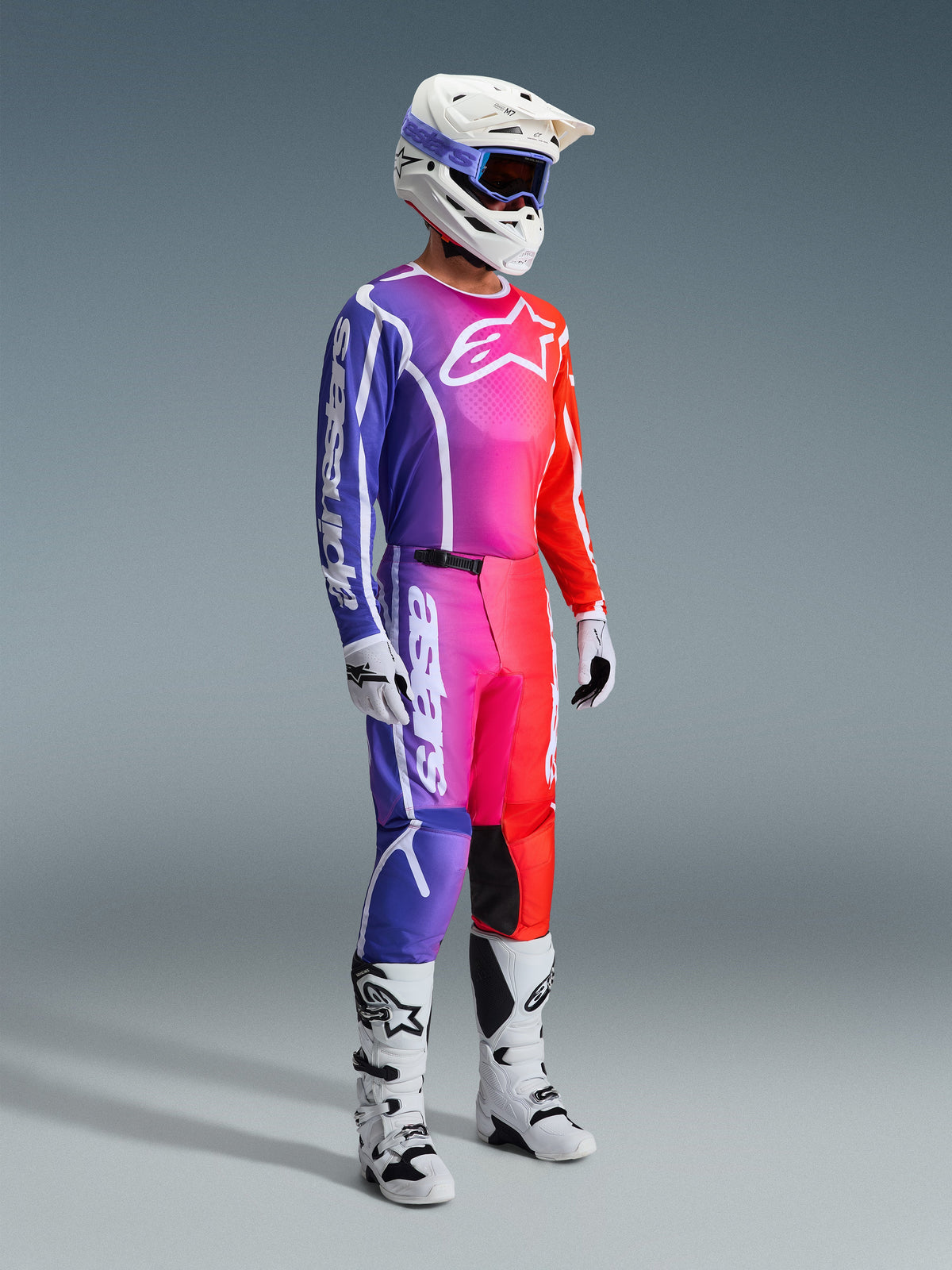 Motocross rider wearing complete racing gear, 2026 Fluid Apex Jersey and Pants with a vibrant purple, pink, and orange gradient, white helmet with blue goggles, white and black boots, and white gloves, coordinated outfit for off-road motocross.