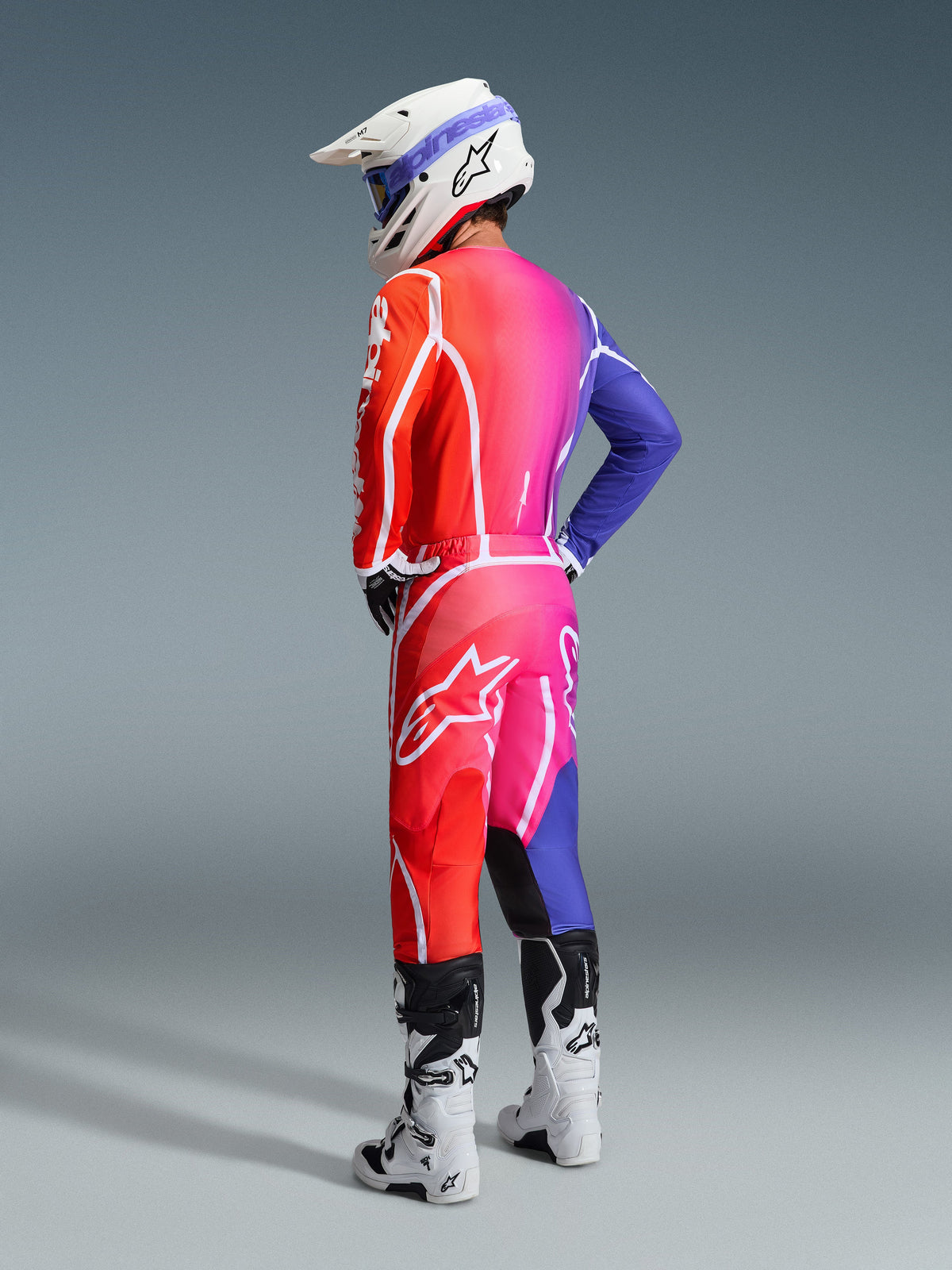 Motocross rider wearing the Alpinestars 2026 Fluid Apex Jersey and matching pants, vibrant red, pink, and blue gradient with white accents, white helmet with blue goggles and white boots, rear view of complete off-road racing gear.