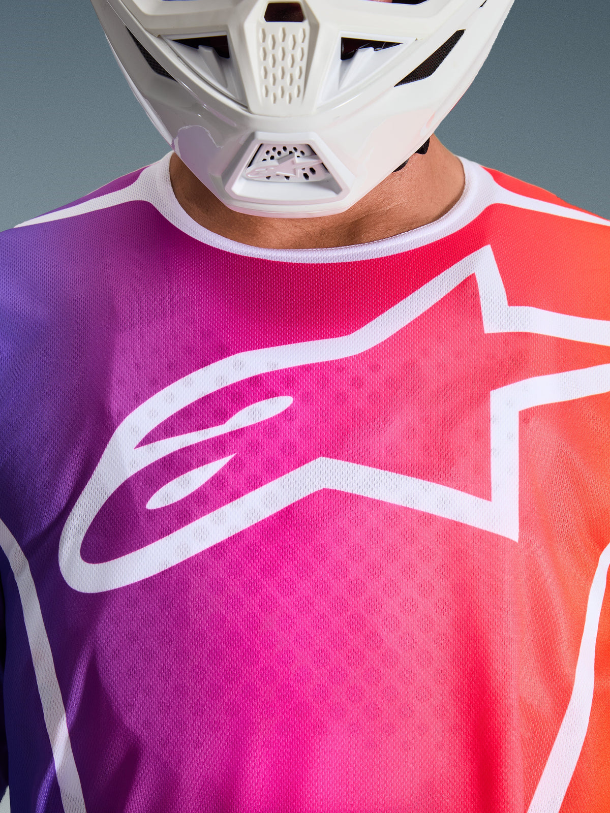 Alpinestars 2026 Fluid Apex Jersey, motocross shirt, paars, roze en oranje kleurverloop, groot wit Alpinestars sterlogo op de borst, close-up weergave met wit kinstuk van de helm