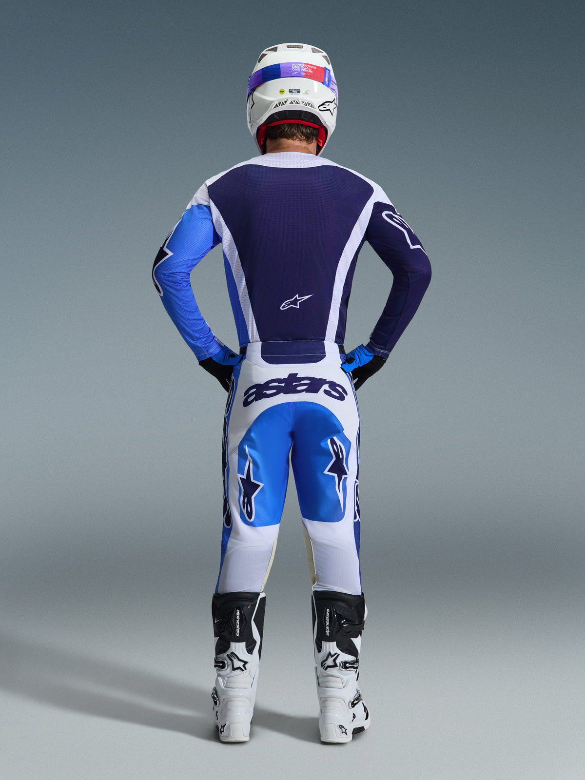 Achteraanzicht van een motorcrosser die de Alpinestars 2026 Racer Air Portl Jersey in Ucla Blue White draagt, gecombineerd met bijpassende witte en blauwe broek en witte laarzen, waarbij de volledige airflow mesh-constructie op de rug voor off-road racen wordt benadrukt.