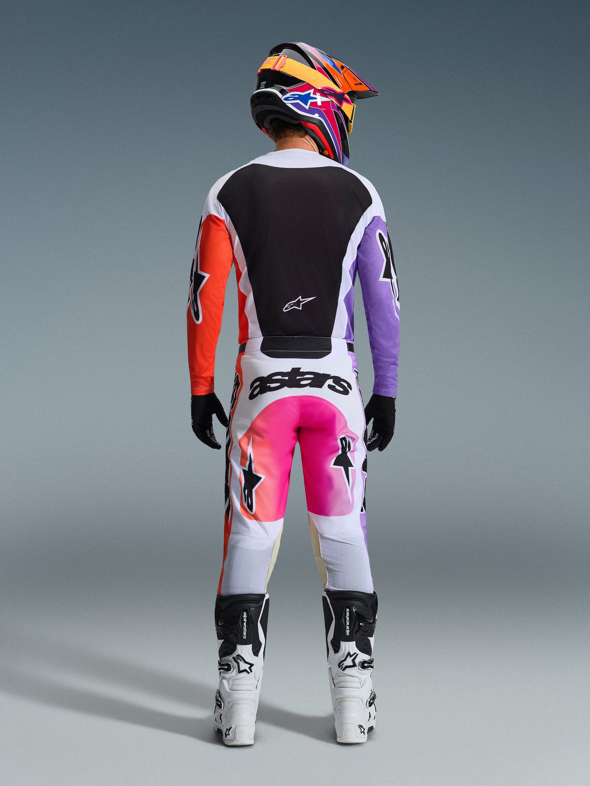 Achteraanzicht van een motorcrosser die de Alpinestars 2026 Racer Air Portl Pants in Multicolor draagt, met witte stof met roze en oranje kleurverlooppanelen en zwarte branding, gecombineerd met een bijpassende zwarte jersey met paarse en oranje mouwen, witte motorlaarzen en een kleurrijke helm.