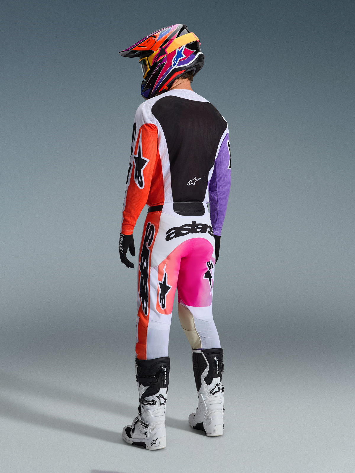 Motocrossrijder van achteren gezien in de Alpinestars 2026 Racer Air Portl broek in Multicolor, met witte, oranje en paarse panelen, neonroze accenten en opvallende zwarte logo's, gecombineerd met een bijpassende jersey, kleurrijke helm met patroon en wit-zwarte laarzen.