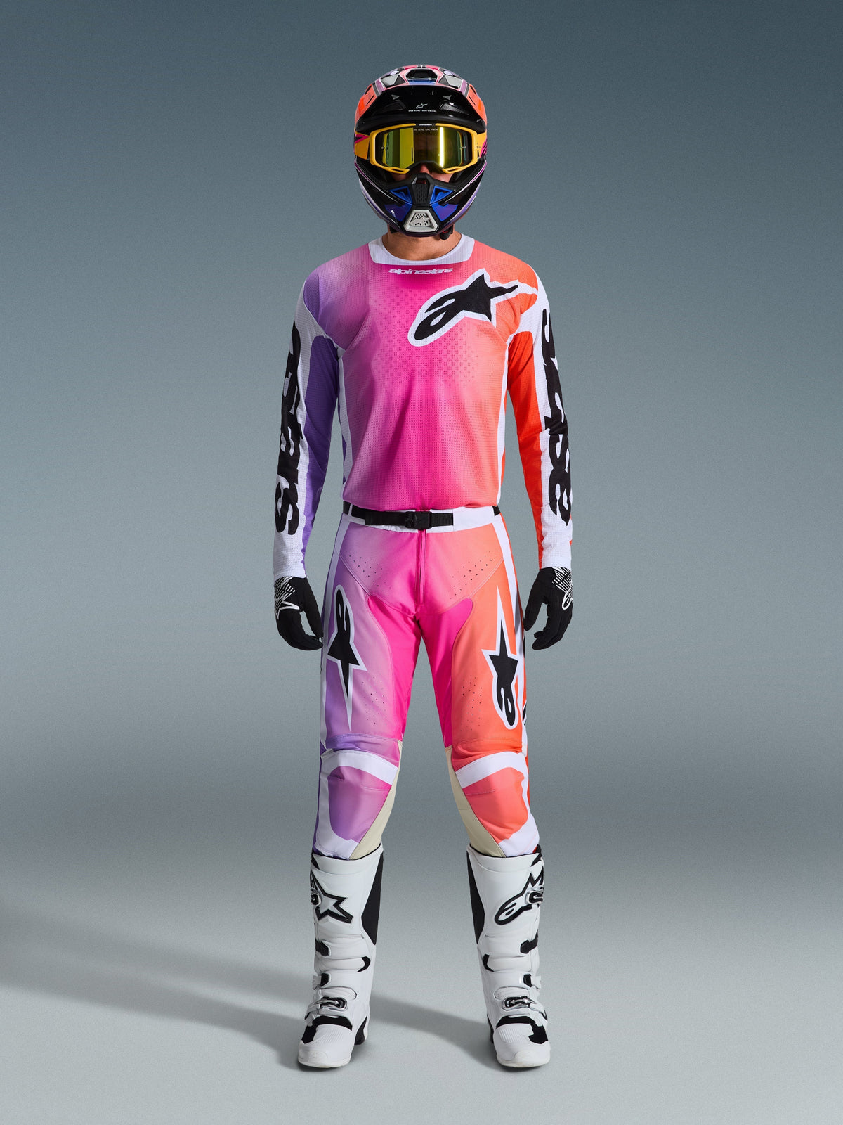 Motorcrosser in volledige Alpinestars 2026 Racer Air Portl race-uitrusting, met een roze, paars en oranje kleurverloop op de broek en jersey, gecombineerd met een bijpassende helm, gele crossbril, witte laarzen en zwarte handschoenen voor een gecoördineerde off-road look
