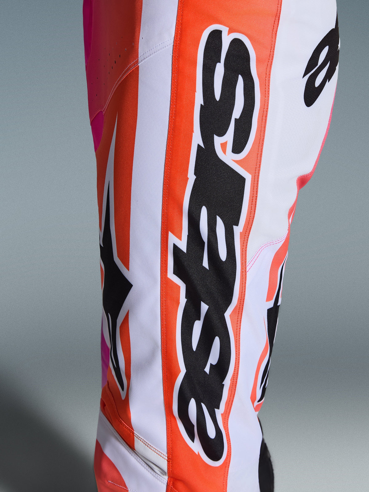 Alpinestars 2026 Racer Air Portl broek, motorcrossbroek, oranje, wit en roze, close-up met grote zwarte Alpinestars-branding en sterlogo, geperforeerde stof voor ventilatie, duurzame off-road racekleding