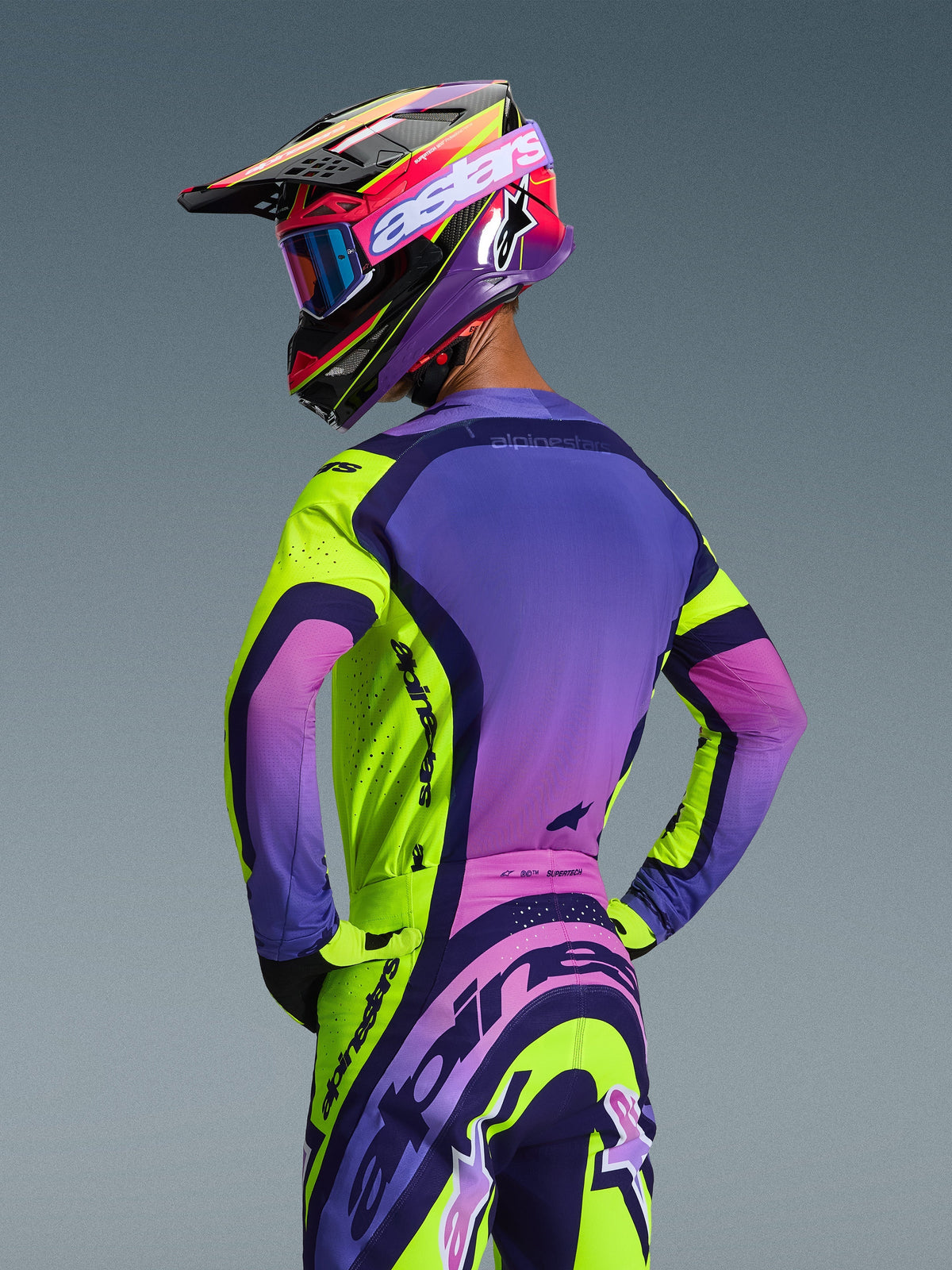 Alpinestars 2026 Supertech Vista Jersey en bijpassende broek, motorcrosser van achteren gezien, kleurstelling Yellow Purple Black met levendige kleurverlopen, gecoördineerde racehelm met blauwe bril en gele handschoenen, professionele off-road race-uitrusting met atletische pasvorm en geïntegreerde branding.