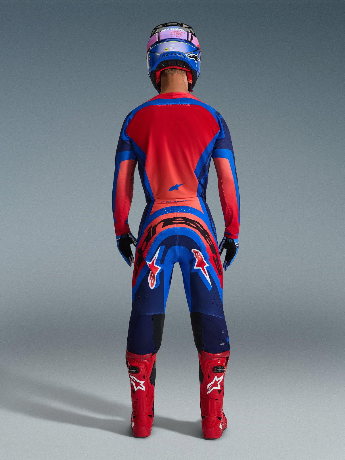 Motocross rider from rear view wearing Alpinestars 2026 Supertech Vista Gear Set in Dark Navy, Hot Coral, and Cobalt, featuring a vibrant red jersey back, bold branding on the pants, coordinated red boots, and a blue and pink helmet.