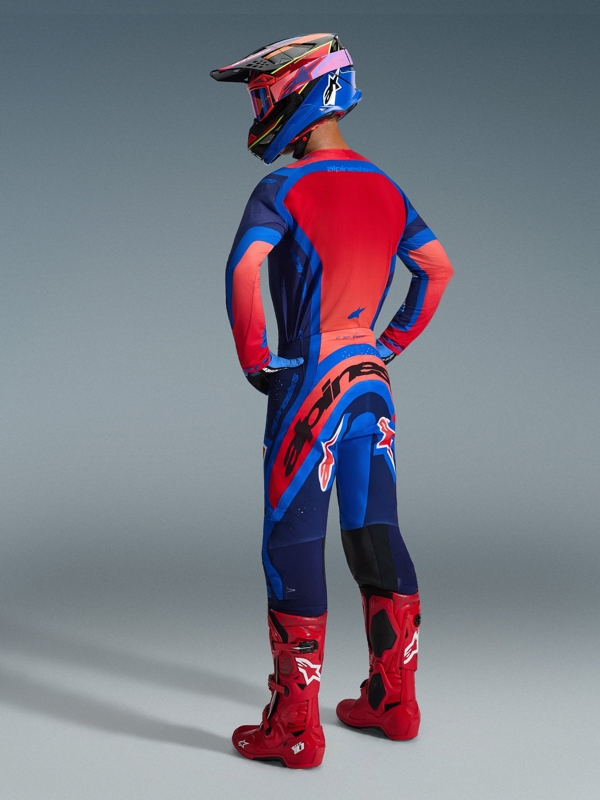 Alpinestars 2026 Supertech Vista Gear Set, rear view of a motocross rider in Dark Navy, Hot Coral, and Cobalt jersey and pants, paired with a multi-colored helmet and bright red motocross boots, professional off-road racing apparel.
