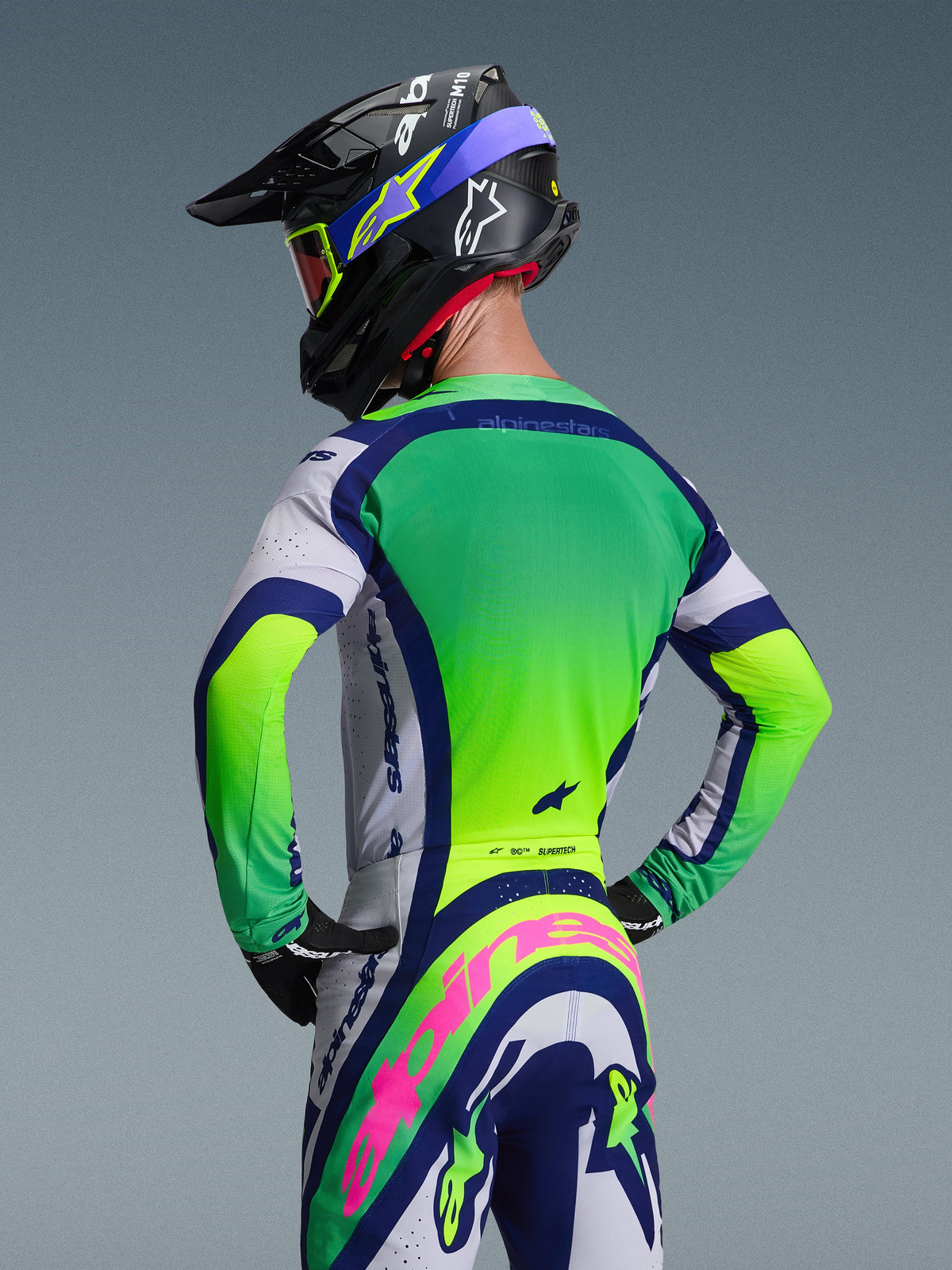 Achteraanzicht van een motorcrosser die de Alpinestars 2026 Supertech Vista Gear Set draagt, met een groen en wit jersey met marineblauwe accenten, een bijpassende broek met een prominent roze Alpinestars-logo, een zwarte racehelm met blauwe graphics en een rood getinte crossbril tegen een neutrale grijze achtergrond.