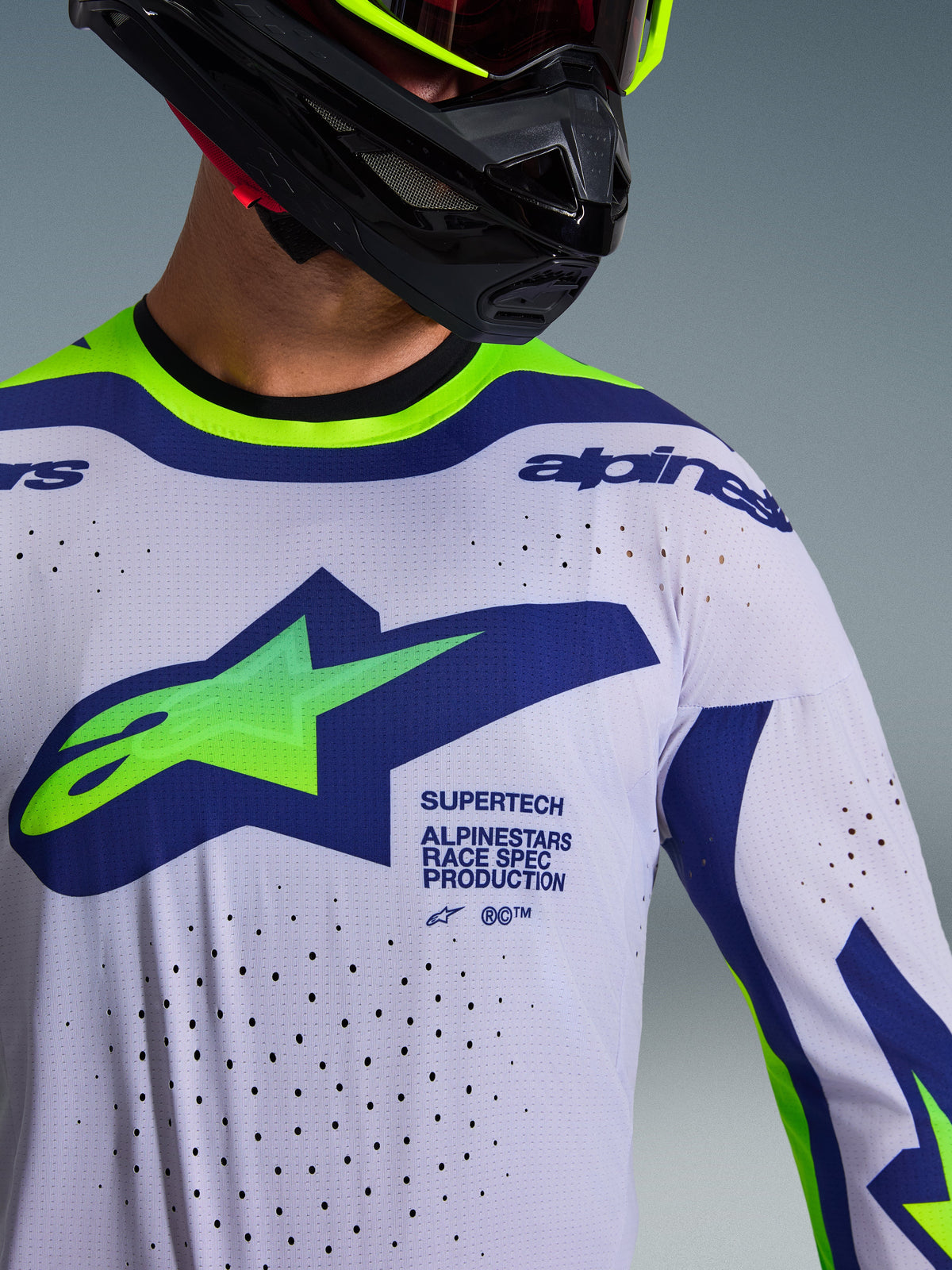 Alpinestars 2026 Supertech Vista Jersey, motorcross jersey, lichtgrijs met geel fluo en blauwe accenten, gedragen door rijder met zwarte helm en neongele crossbril, groot groen en blauw Astars-logo, lasergesneden ventilatiegaten, atletische racepasvorm
