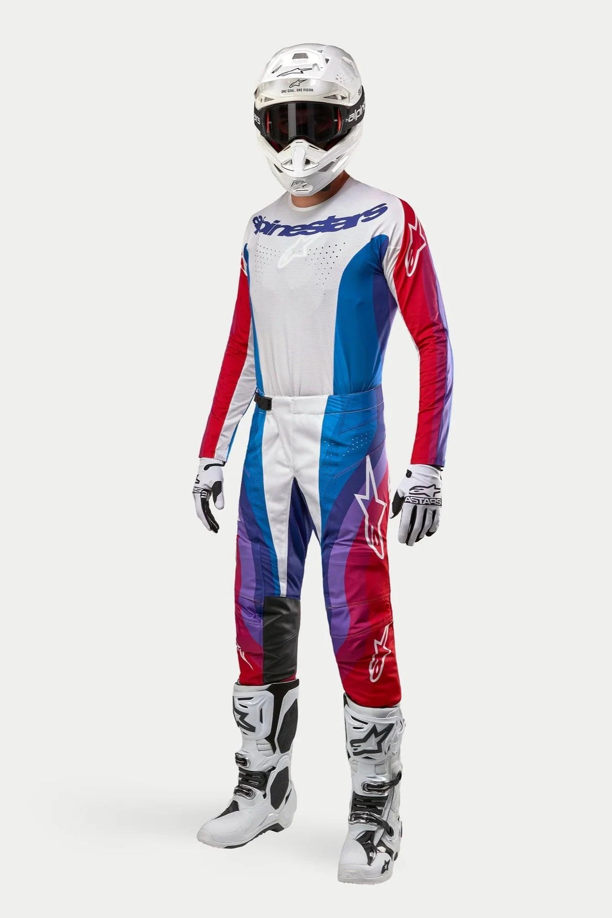 Motorcrosser in volledige Alpinestars-uitrusting, 2024 Techstar Pneuma broek in Blue Mars Red White, bijpassende jersey met lange mouwen, witte helm met donkere bril en wit-zwarte laarzen, gecoördineerde outfit voor off-road racing.