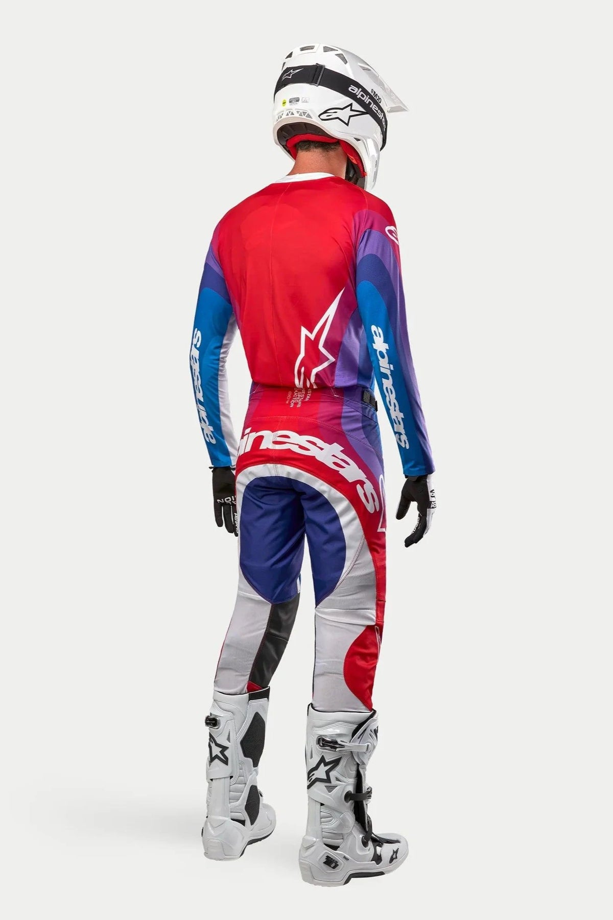 Alpinestars 2024 Techstar Pneuma jersey en broek, motorcrosser van achteren gezien, Blu Mars Red White kleurstelling met rood, blauw en paars kleurverloop, witte logo's, gecombineerd met witte laarzen en een witte helm, gecoördineerde off-road race-uitrusting.