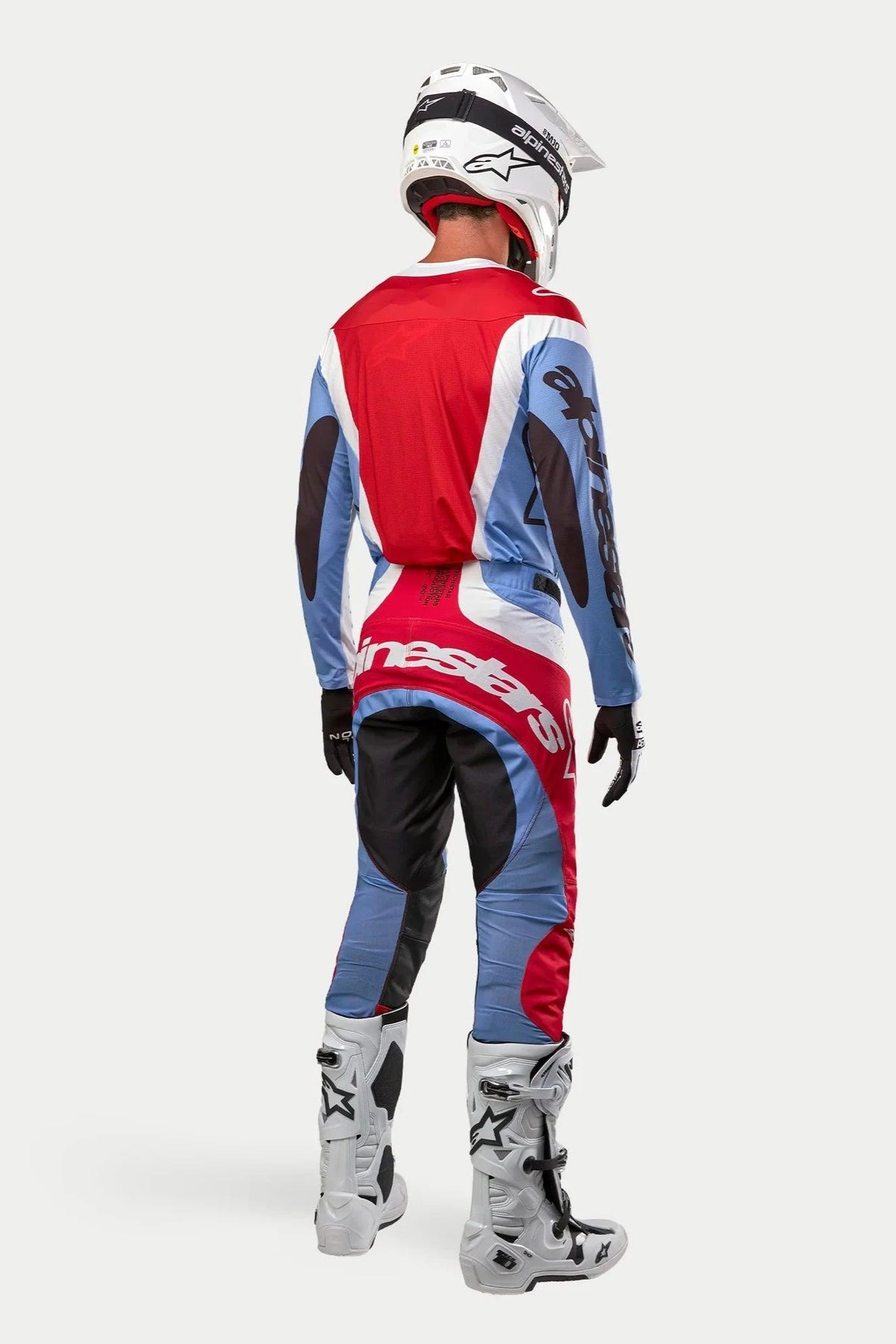 Alpinestars 2024 Techstar Ocuri motorcross outfit van achteren gezien, jersey en broek in lichtblauw, Mars-rood en wit, witte helm met zwarte graphics, witte en grijze off-road laarzen, zwarte handschoenen, complete race-uitrusting