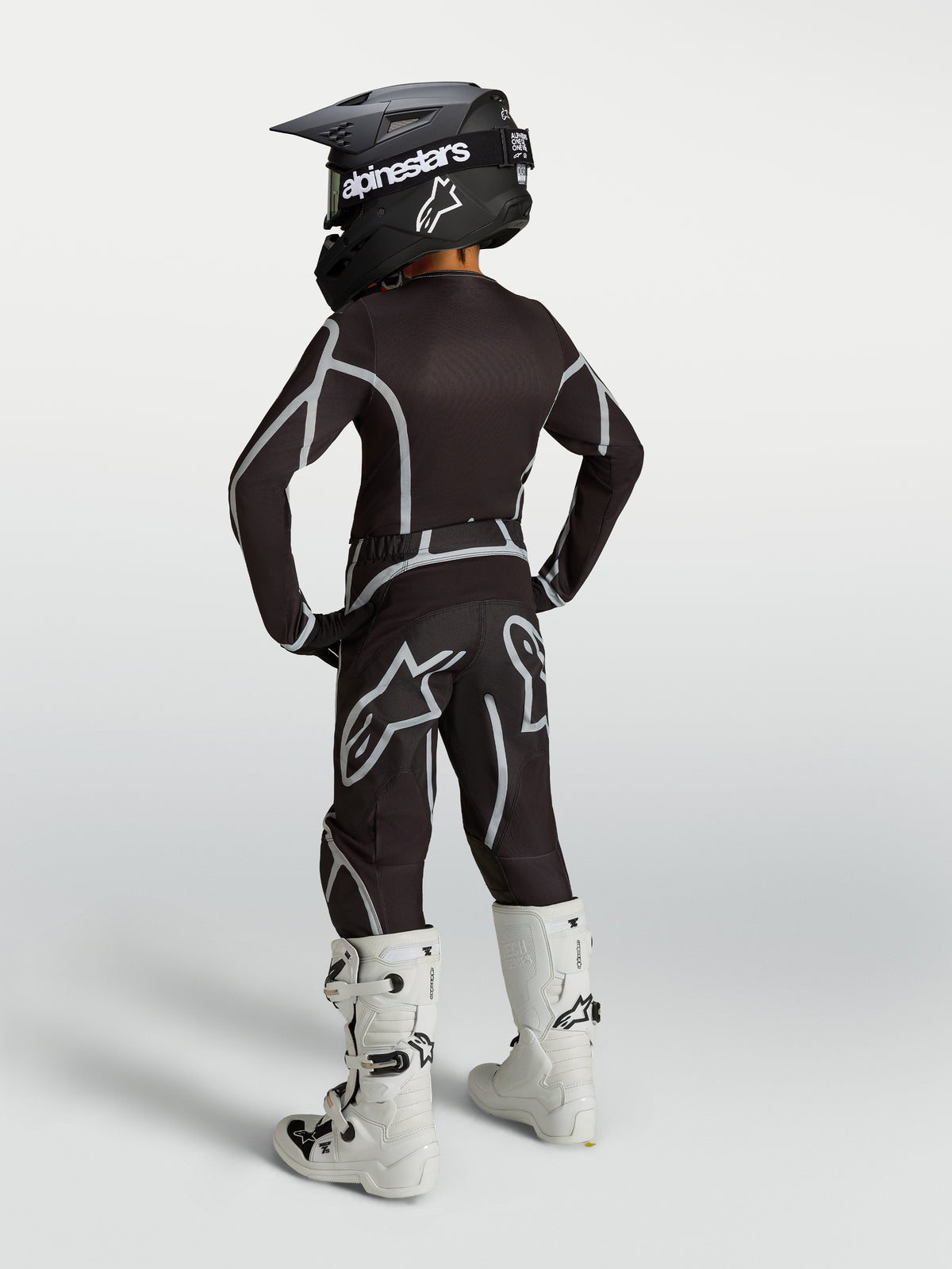Alpinestars 2027 Jeugd Fluid Apex V2 gear set voor motorcross, zwart en zilver, achteraanzicht met witte motorlaarzen.