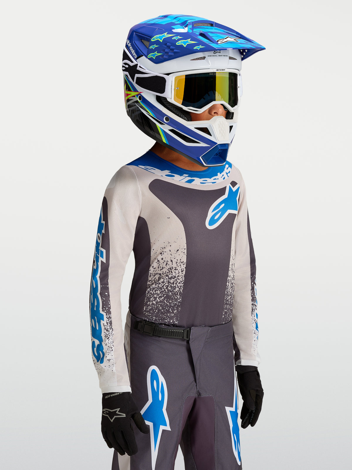 Alpinestars Jeugd Fluid Drag Jersey 2027, motorcrossjersey voor kinderen, grijs en blauw, lichtgewicht ademende constructie, gedragen door een model met een motorcrosshelm en handschoenen, professionele off-road race-uitrusting.