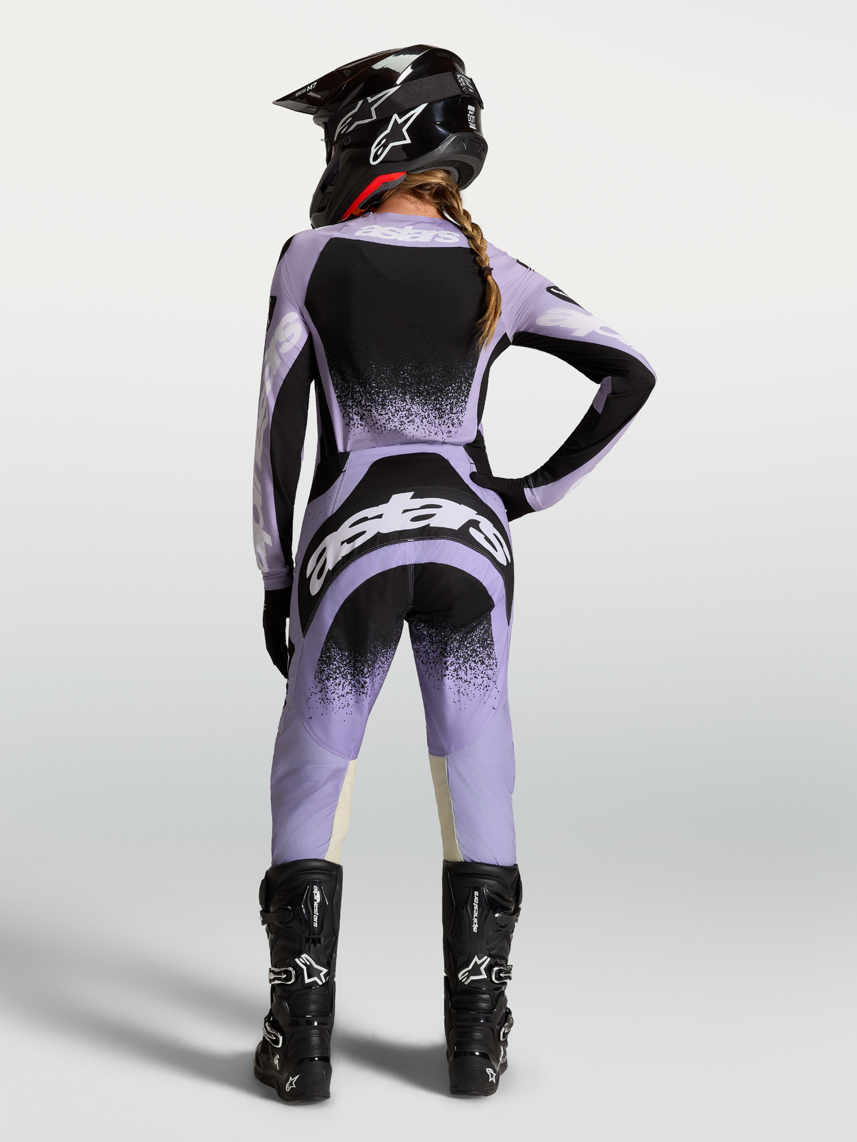Alpinestars Stella Techstar Swan broek, paarse en zwarte dames motorcrossbroek voor racing, ergonomische pasvorm, achteraanzicht van model in complete MX-uitrusting met zwarte motorlaarzen en crosshelm.