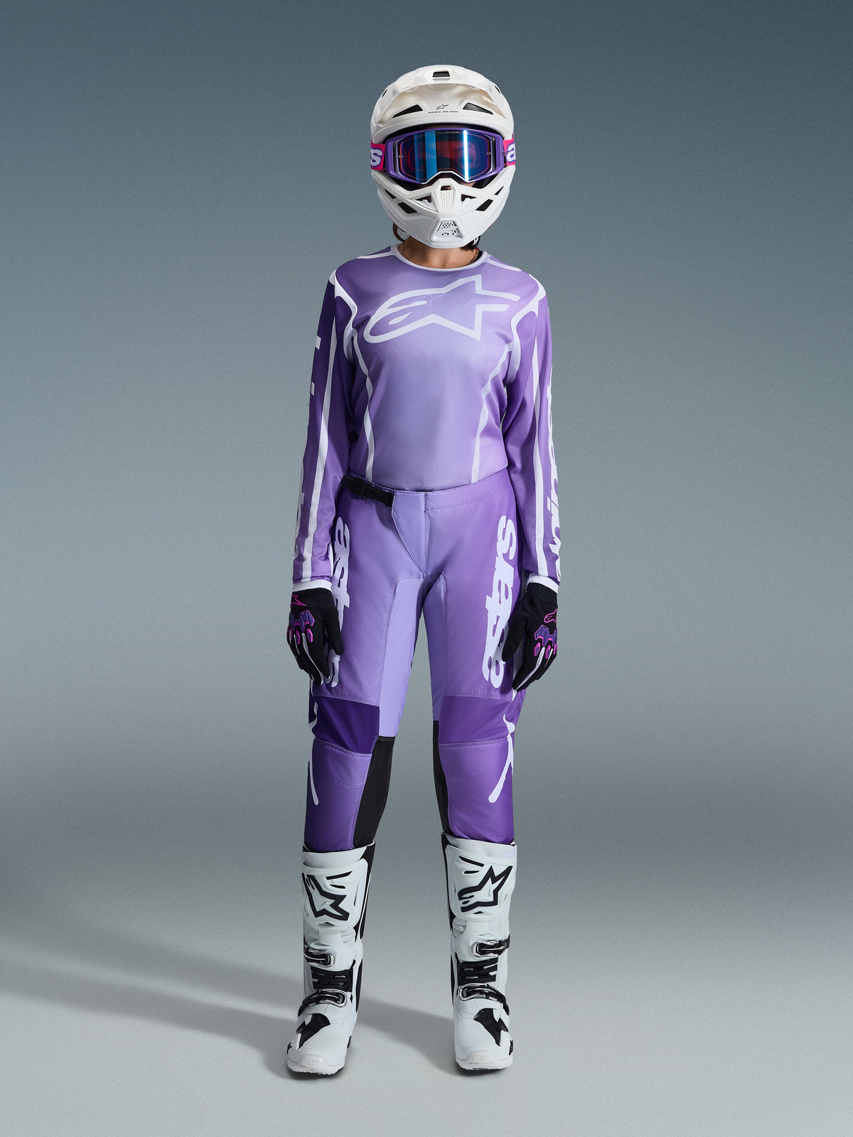 Motorcrossrijder in de Alpinestars 2026 Stella Fluid Apex Gear Set, paarse jersey met wit logo, bijpassende paarse broek met witte branding, witte helm met roze bril, wit-zwarte laarzen, gecoördineerde dames motorcross outfit voor off-road racing