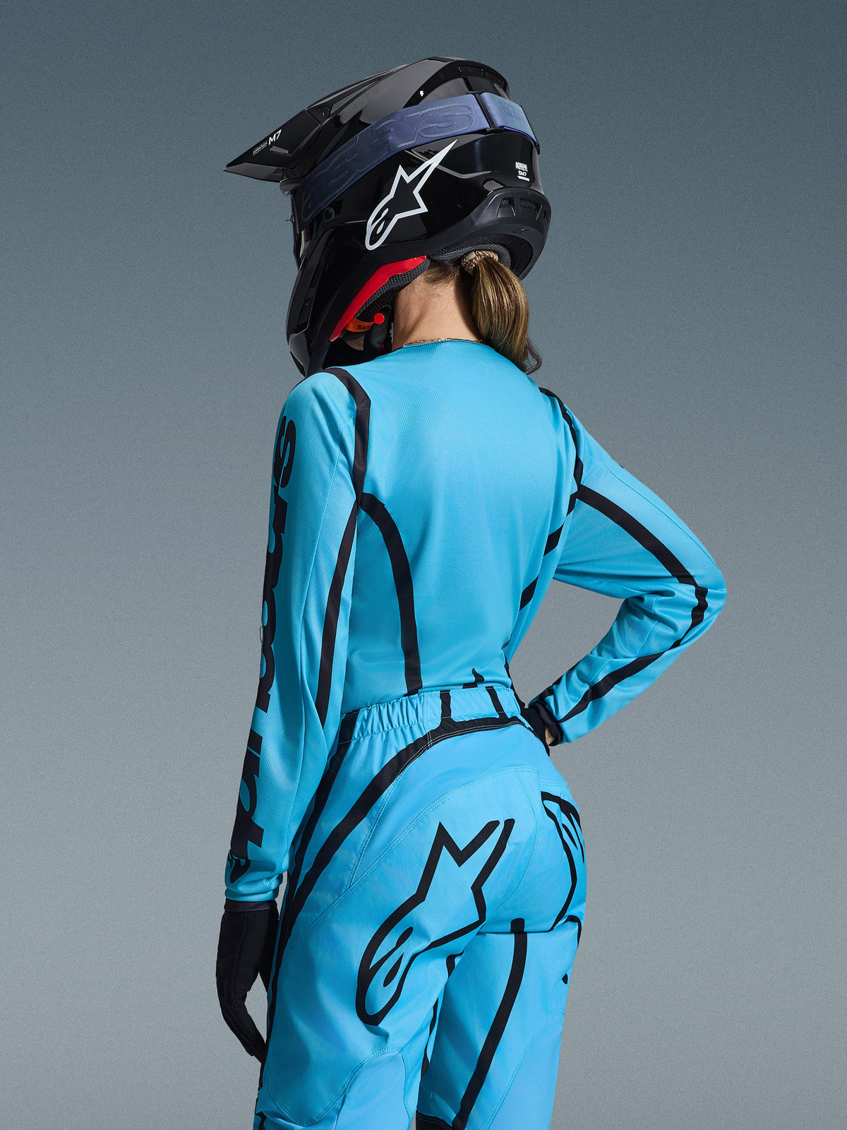 Alpinestars 2026 Stella Fluid Apex Gear Set, lichtblauw en zwart, achteraanzicht van een vrouwelijke rijder die een motorcross jersey en bijbehorende broek draagt, zwarte Alpinestars-helm met wit logo, technische off-road racekleding voor dames.