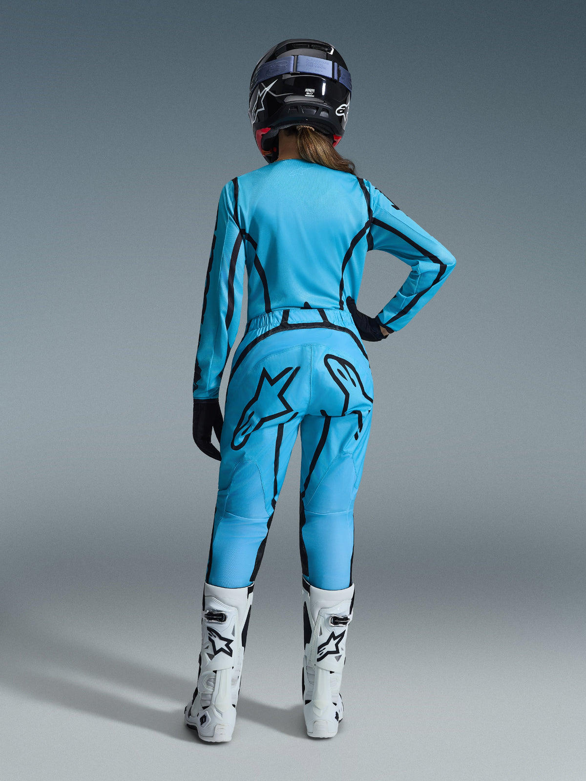 Vrouwelijke motorcrossrijder van achteren, gehuld in de Alpinestars 2026 Woman Stella Fluid Apex Gear Set in helderblauw met zwarte grafische lijnaccenten, voorzien van een groot zwart ster-logo op de broek, gecombineerd met witte en zwarte laarzen en een zwarte helm.