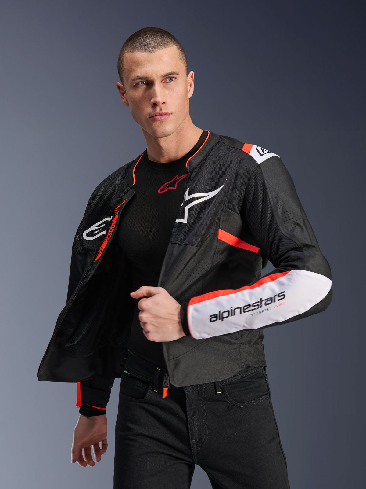 Alpinestars T-SPS Air V2 Jas, zwarte textiele motorjas met witte en fluorescerend rode accenten, voorzien van mesh-panelen voor luchtstroom, wit Alpinestars-logo op de borst en merknaam op de mouw, mannelijk model draagt de jas open over een zwarte basislaag
