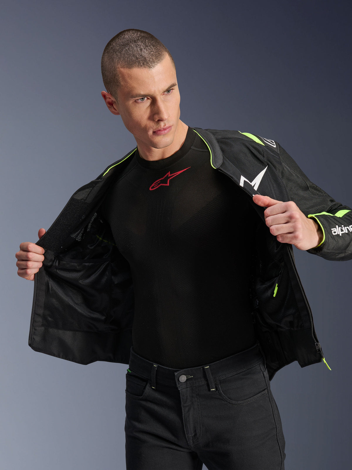Mannelijk model dat de Alpinestars T-Sps Air V2 Jas in Black Yellow Fluo draagt, open getoond om de interne mesh-constructie en zakken te benadrukken, gecombineerd met een zwarte Alpinestars base layer en zwarte broek, stedelijke motorrijstijl.