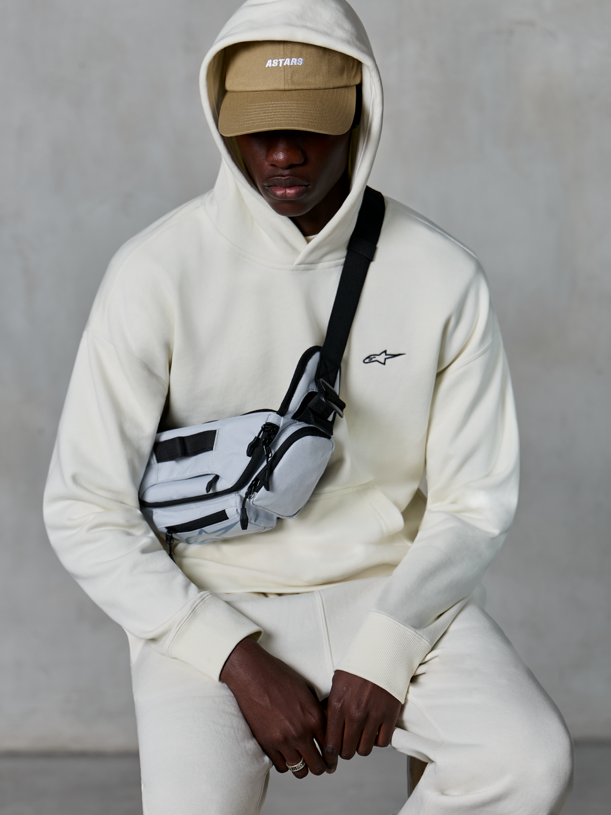 Alpinestars Essentials Ageless Hoodie, casual streetwear, Off White, small black embroidered logo on chest, hood worn over a tan cap with ASTARS branding, styled with a light grey cross-body bag, premium lifestyle apparel