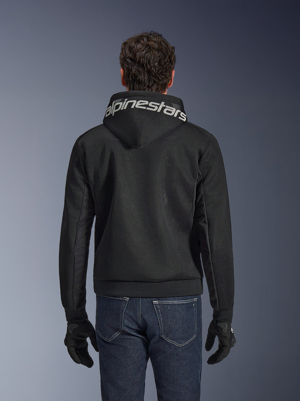 Alpinestars Chrome Superairflow Hoodie, zwarte motorhoodie, achteraanzicht met opvallende witte Alpinestars-branding op de capuchon, 3D technische mesh-constructie, gedragen met donkerblauwe jeans en zwarte handschoenen, geventileerde stedelijke motorkleding