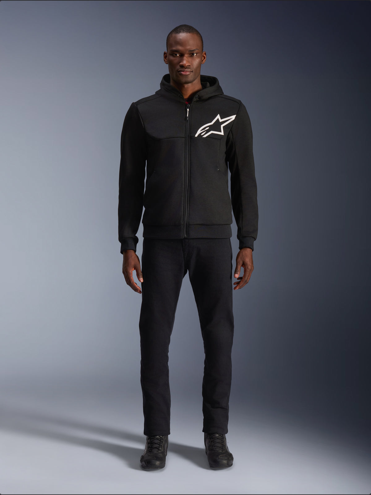 Alpinestars Chrome V2 Sport Hoodie, zwart-witte motorhoodie met een groot wit Alpinestars-logo op de borst, ritssluiting en capuchon, gedragen door een model met zwarte broek en laarzen, urban-stijl beschermende motorkleding.