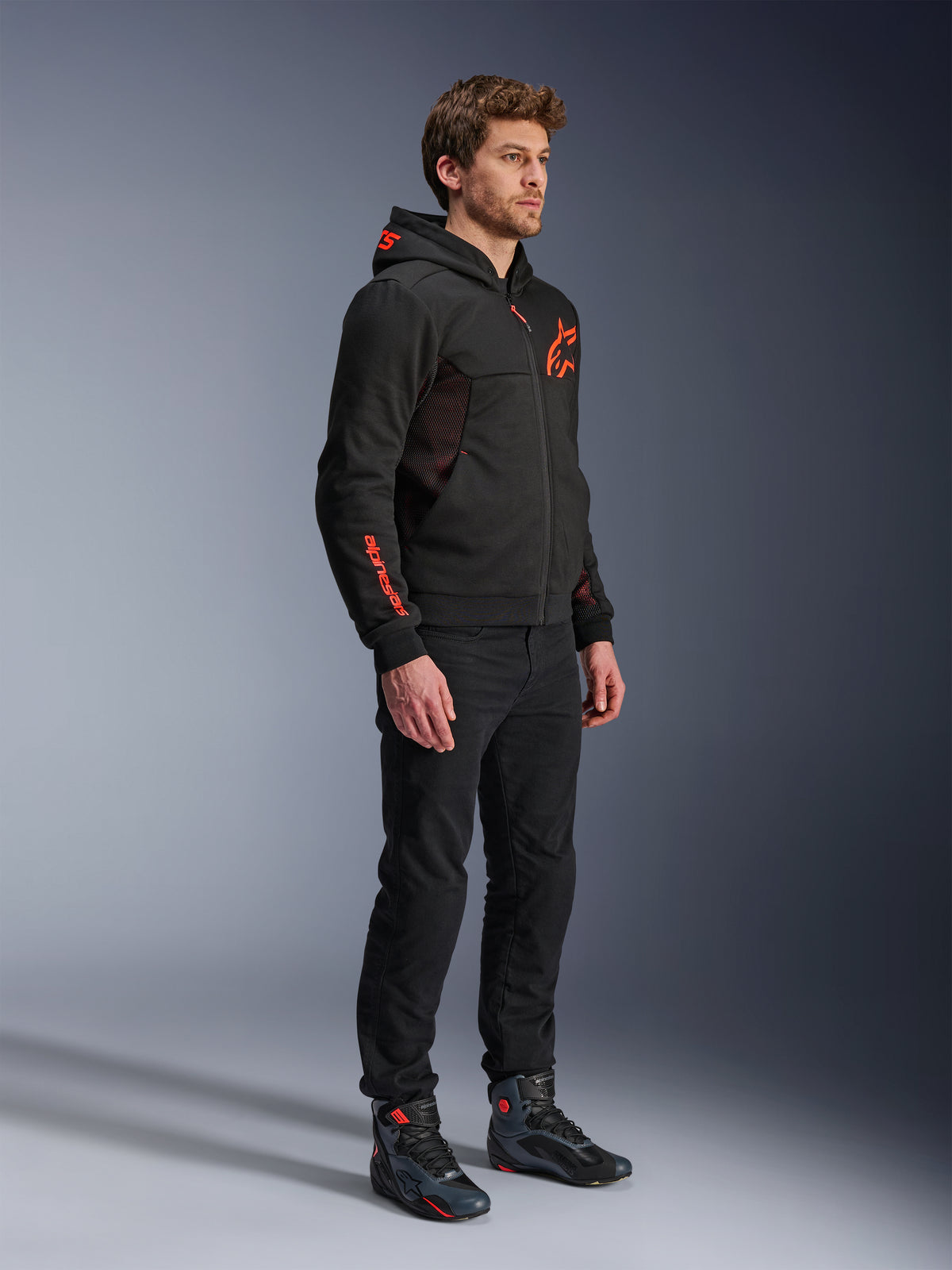 Alpinestars Chrome Air Sport Hoodie, Black Red Fluo, urban motorjas met tech fleece, gedragen met zwarte broek en motorlaarzen, voorzien van mesh ventilatiepanelen en rode fluorescerende logo's op de borst en mouw.