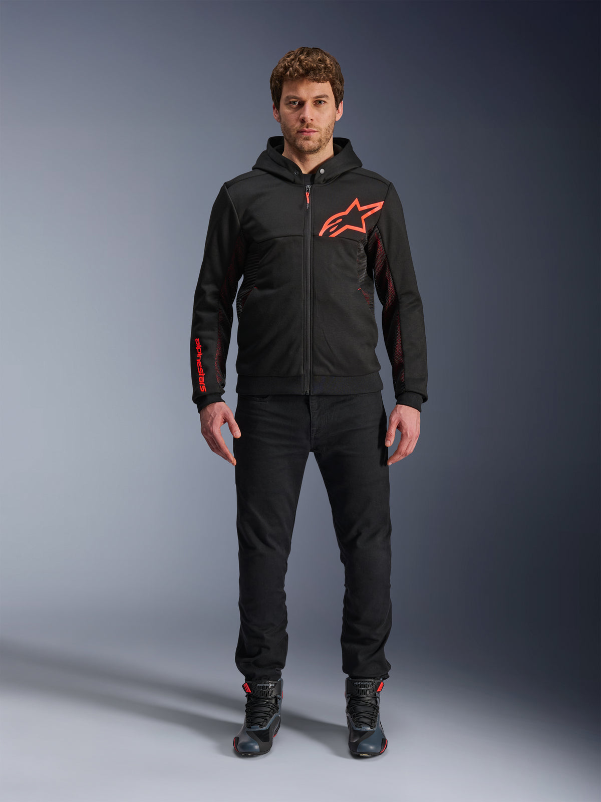 Alpinestars Chrome Air Sport Hoodie, zwart en fluorrood, urban motorhoodie met tech fleece constructie, voorzien van een groot rood Astars-logo op de borst en merknaam op de mouw, gedragen door een model met een zwarte broek en motorschoenen