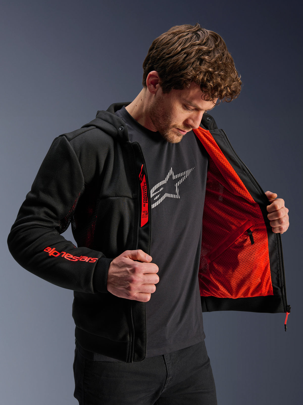 Alpinestars Chrome Air Sport Hoodie in Black Red Fluo, mannelijk model opent de jas om de felrode mesh voering te tonen, Alpinestars-branding op de rechtermouw en borstrits, casual stedelijke motorjas ontworpen voor optimale luchtstroom.