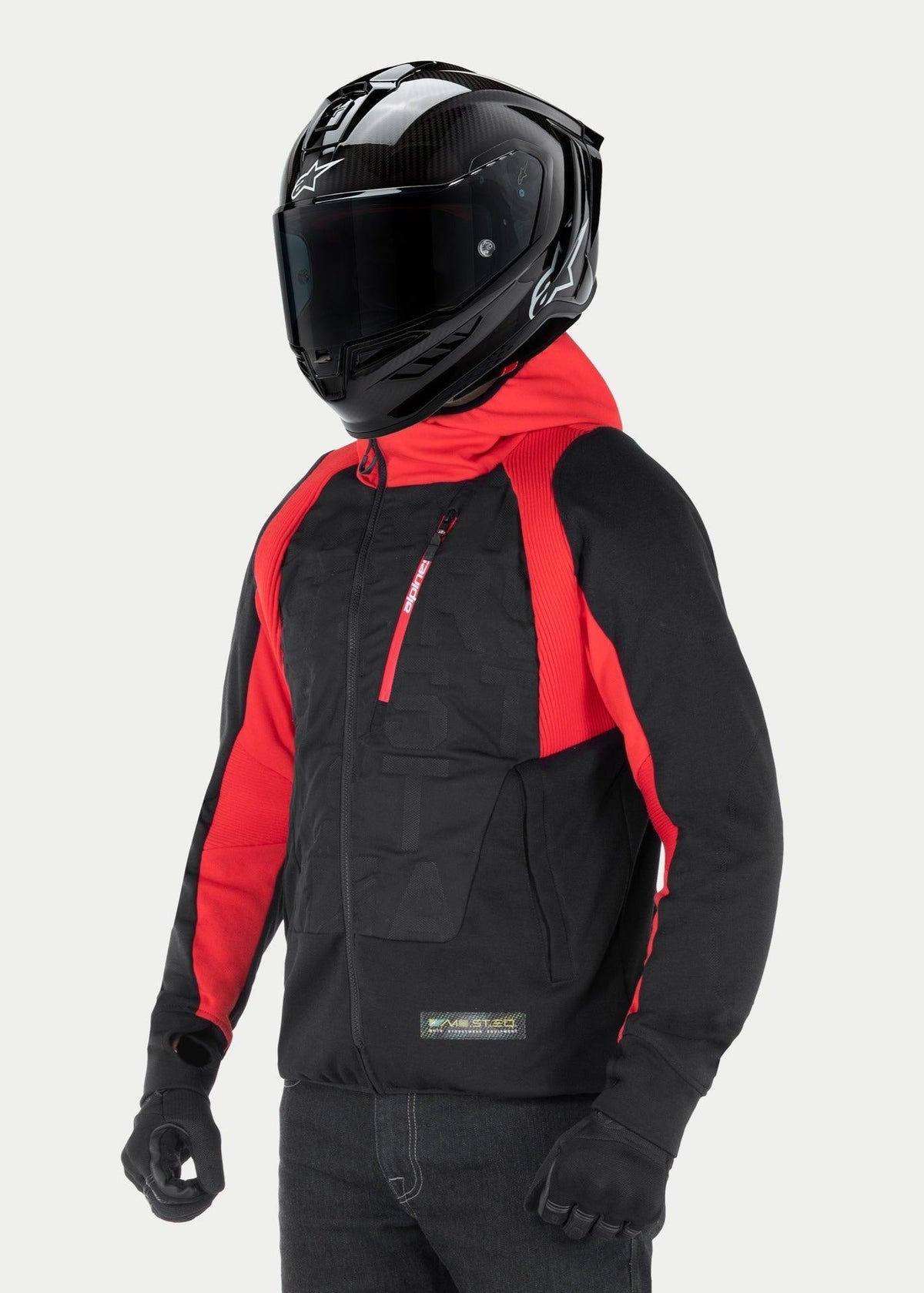 Alpinestars MO.ST.EQ Hybrid Hooded Jacket, urban motorhoodie, zwart en rood, voorzien van een schuine rits aan de voorzijde en geribbelde schouderpanelen, gedragen met een zwarte integraalhelm en zwarte handschoenen, eigentijdse streetwear-stijl