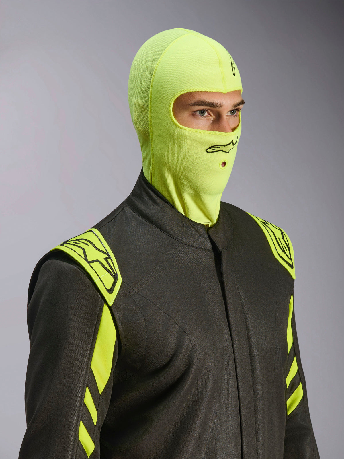 Mannelijk model draagt Alpinestars ZX Evo V3 bivakmuts in Yellow Fluo, vuurbestendige race-bivakmuts, gecombineerd met een donkergrijs autoracepak met neongele accenten, voorzien van zwart Alpinestars-logo en mondopening voor hydratatieslang, professionele beschermende autosportuitrusting