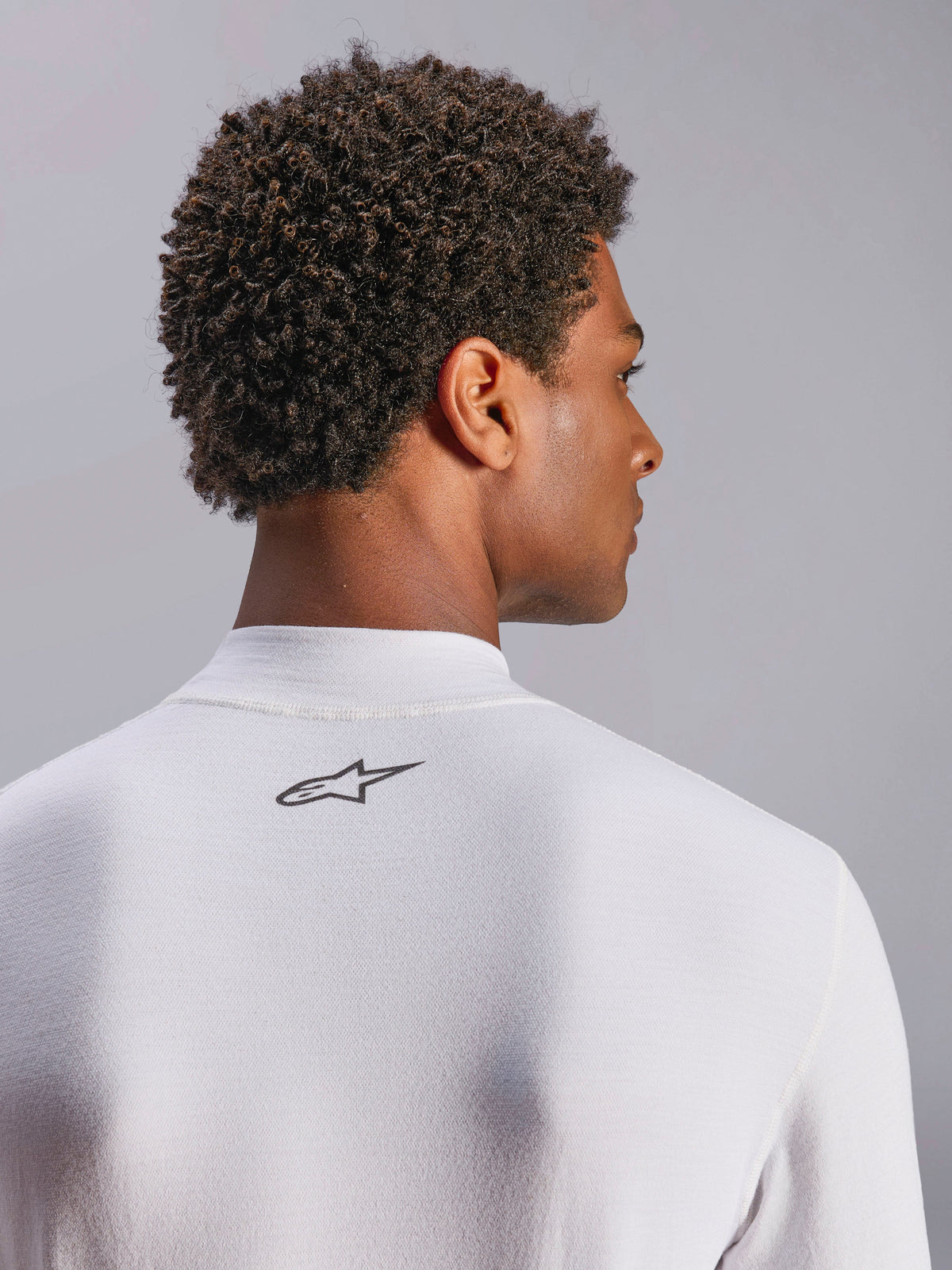Alpinestars ZX Evo V3 Top, baselayer voor autoracen met lange mouwen, wit, close-up van het nekgedeelte op de rug met zwart Alpinestars-logo, technische vlamvertragende stof, gedragen door model met donker krullend haar