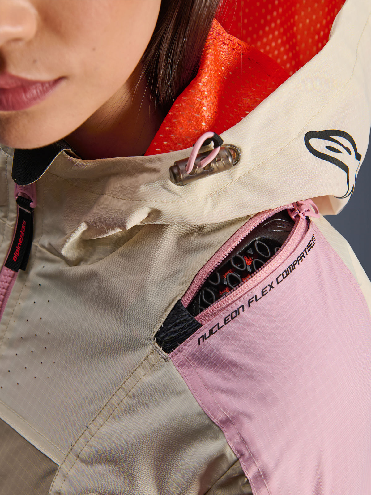 Close-up van de Alpinestars Molly Air Jacket in Sand Pink Almond, met aandacht voor het schoudercompartiment met ritssluiting voor Nucleon Flex-protectoren, de feloranje mesh-voering van de capuchon en de duurzame ripstop-stofconstructie.
