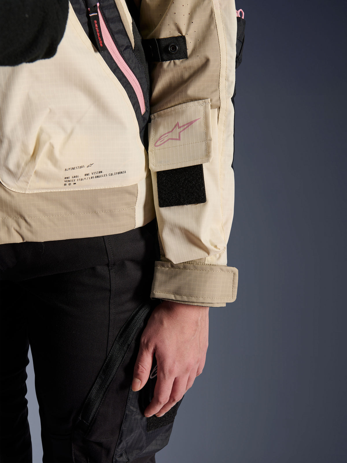Alpinestars Molly Air Jacket, urban motorjas voor dames, Sand Pink Almond, close-up zijaanzicht met mouwzak met roze logo, zwarte klittenbandpatch en merkmissietekst op de onderrug, lichtgewicht ripstop-materiaal