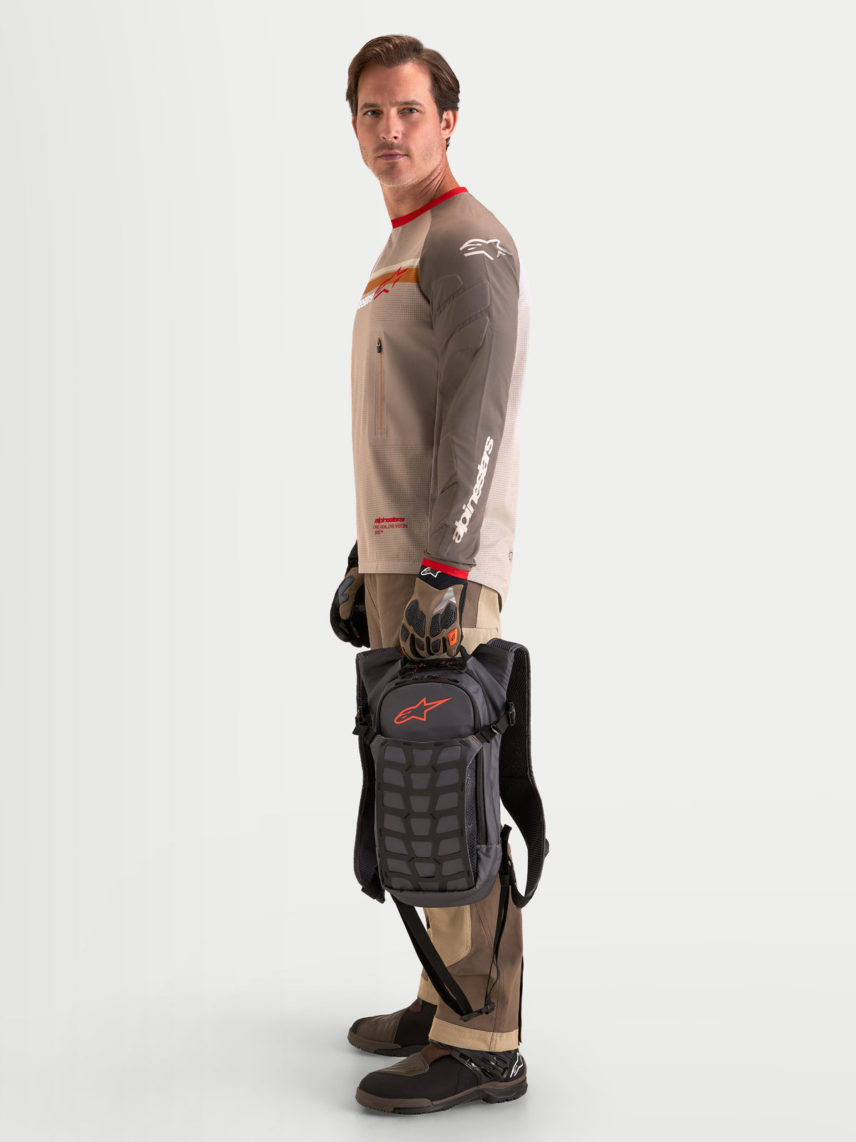 Alpinestars AT-9 rugzak, motocross rugzak in walnootbruin en donkergrijs, zijaanzicht van een model dat staat en de lichte textieltas bij het handvat vasthoudt