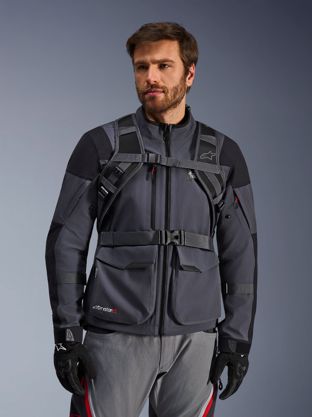 Alpinestars AT-15 Back Pack harness system, Black Deep Gray, front view worn by a rider over a gray motorcycle jacket, featuring magnetic chest lock and secure waist strap, paired with black gloves for adventure motorcycling