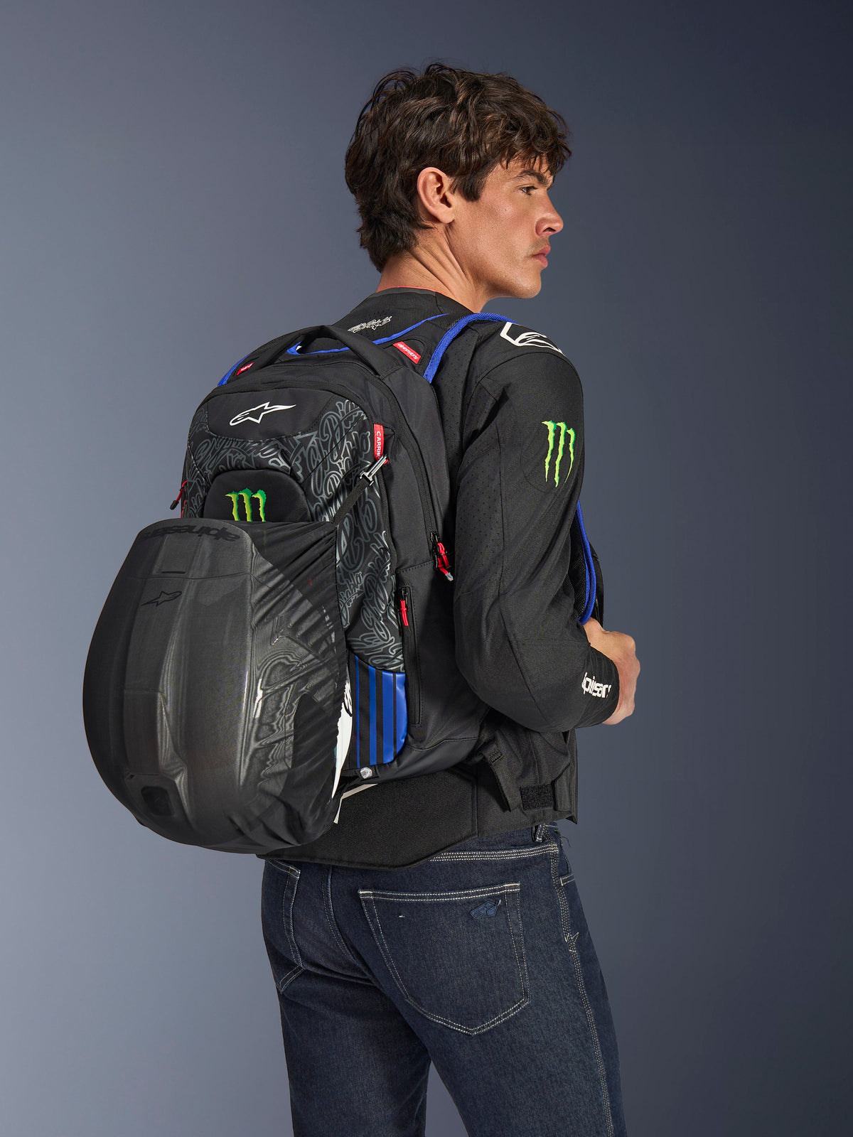 Alpinestars FQ20 City Hunter V2 Monster Backpack, urban motorcycle backpack in Black with Red Fluo, Blue, and Green accents, worn by a model with a black helmet secured in the rear mesh carrier, styled with a black motorcycle jacket and blue jeans.