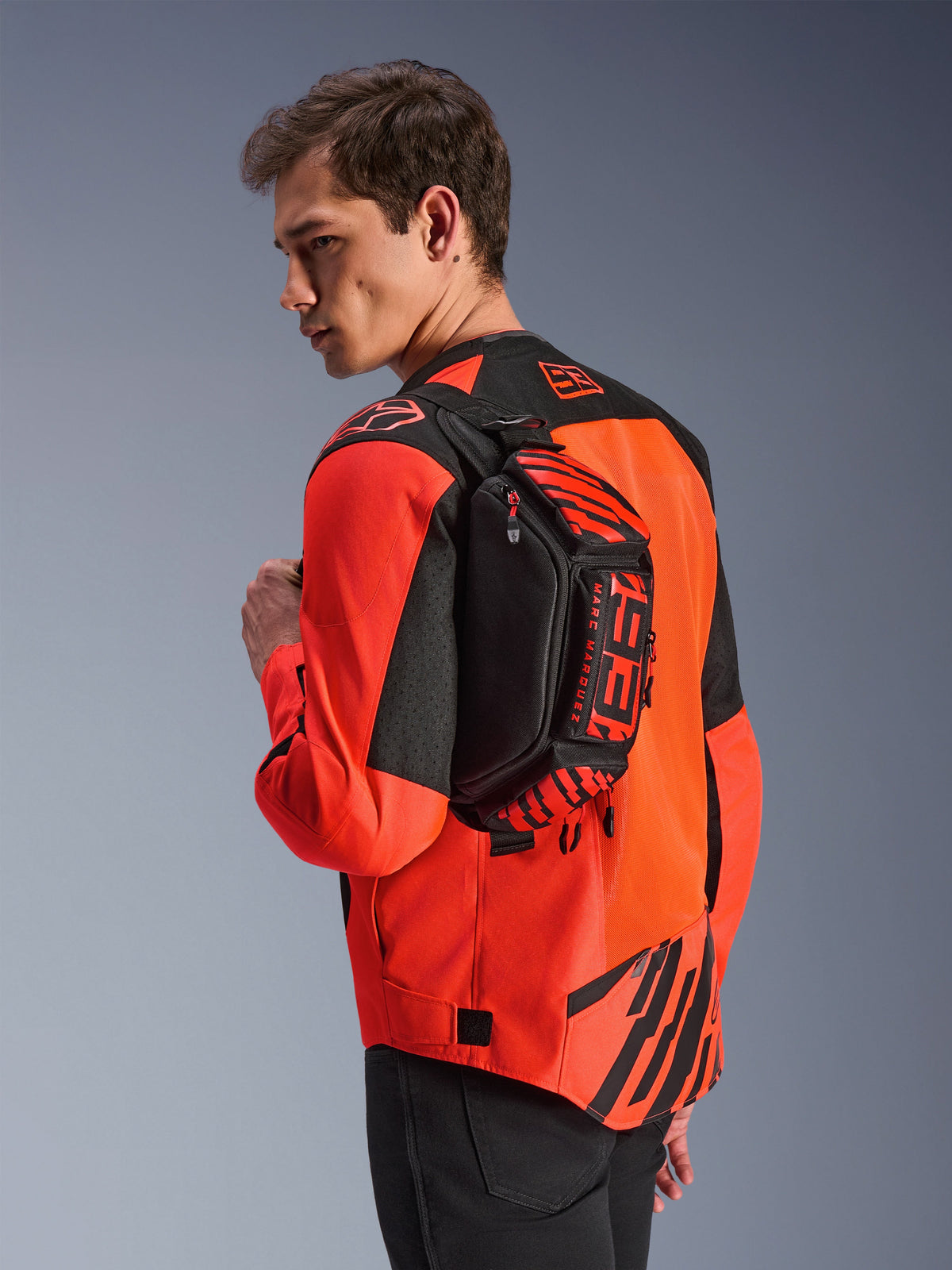 Alpinestars MM93 Waist Bag, heuptas, zwart met Red Fluo en Bright Red, door een model over de rug gedragen over een rood-zwarte motorjas, voorzien van opvallende MM93-branding en Marc Marquez-graphics