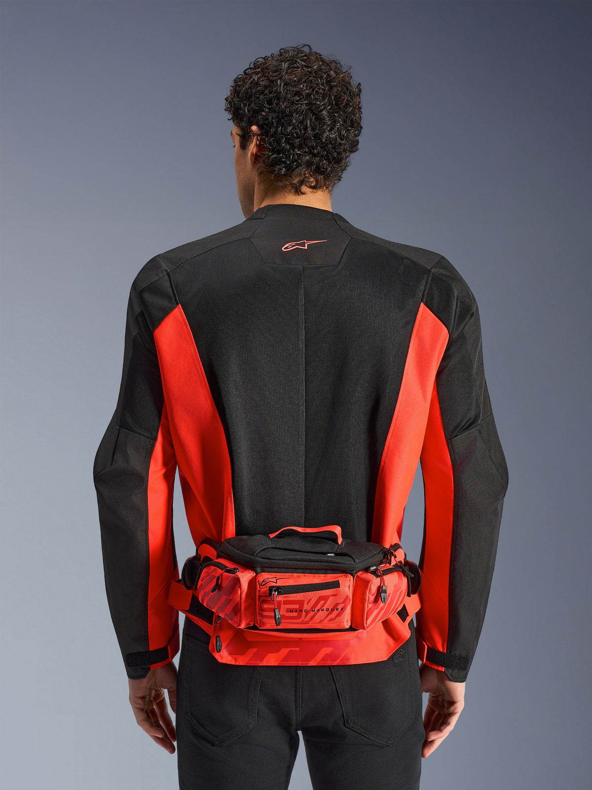 Alpinestars MM93 heuptas, rood en zwart, gedragen over een zwart en rood motorjack, voorzien van Marc Marquez 93-branding, verstelbare riem en vakken met ritssluiting, functioneel op de racerij geïnspireerd accessoire