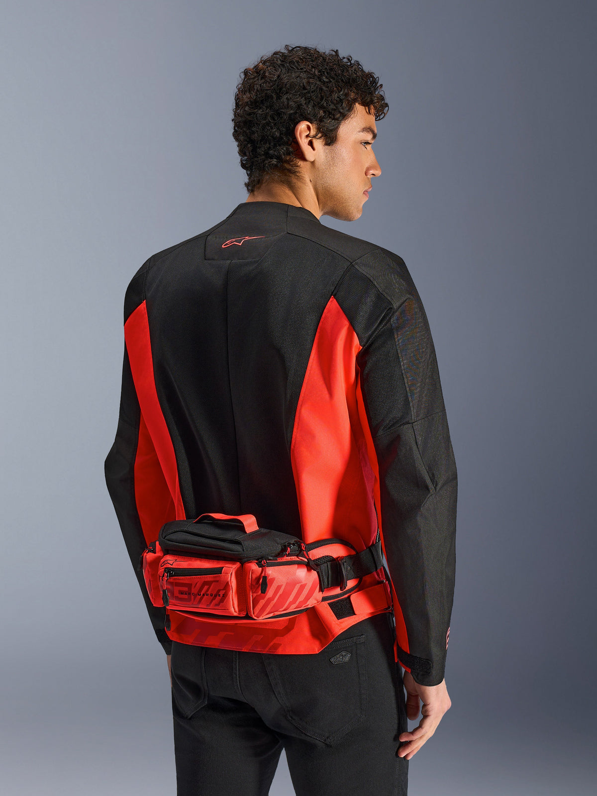 Alpinestars MM93 Waist Bag, motorheuptas, felrood en zwart, gedragen over een bijpassende zwart-rode motorjas, met ritsvakken en Marc Marquez MM93-branding, duurzame textielconstructie voor motorrijden