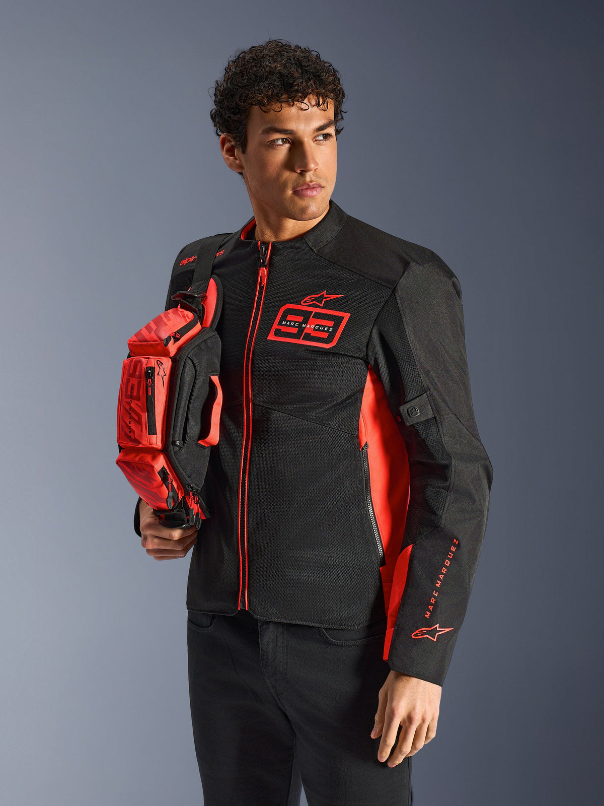 Alpinestars MM93 heuptas, fluorescerend rood en zwart, vastgehouden door een model in een bijpassende zwart-rode motorjas, voorzien van meerdere vakken met ritssluiting en Marc Marquez-branding, praktische uitrusting voor sportief rijden