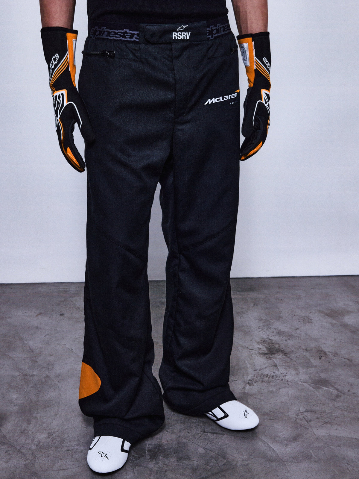 Alpinestars RSRV Arena Suit racingbroek, zwart met oranje details, McLaren-logo's, premium motorsport pantalon, gedragen met race-uitrusting, getextureerde grijze ondergrond