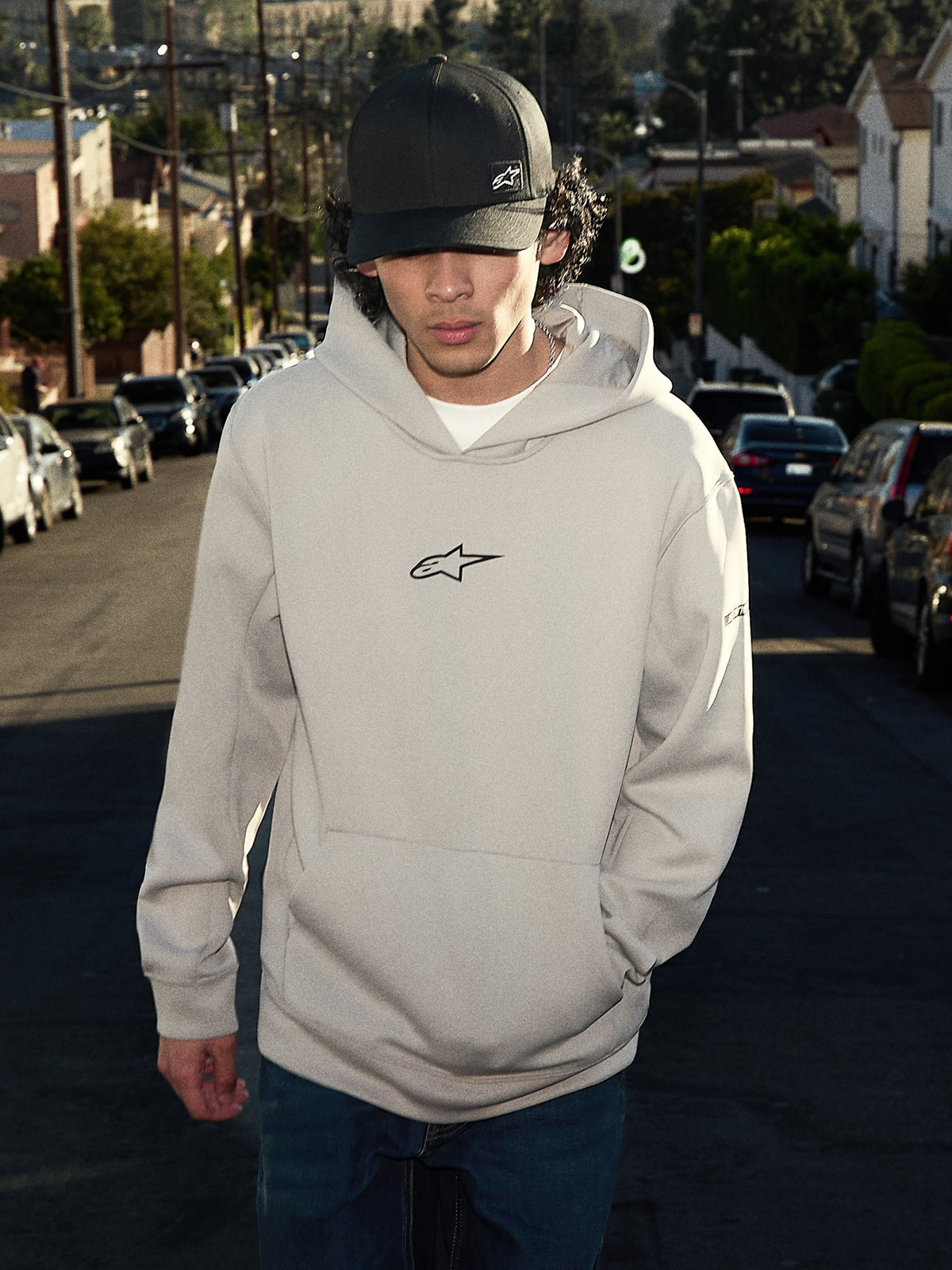 Alpinestars Insight Hoodie, casual lifestyle streetwear, Cement gray with black Astars logo on chest, pullover style with kangaroo pocket, worn with a black cap on a residential street, modern motorsport-inspired design