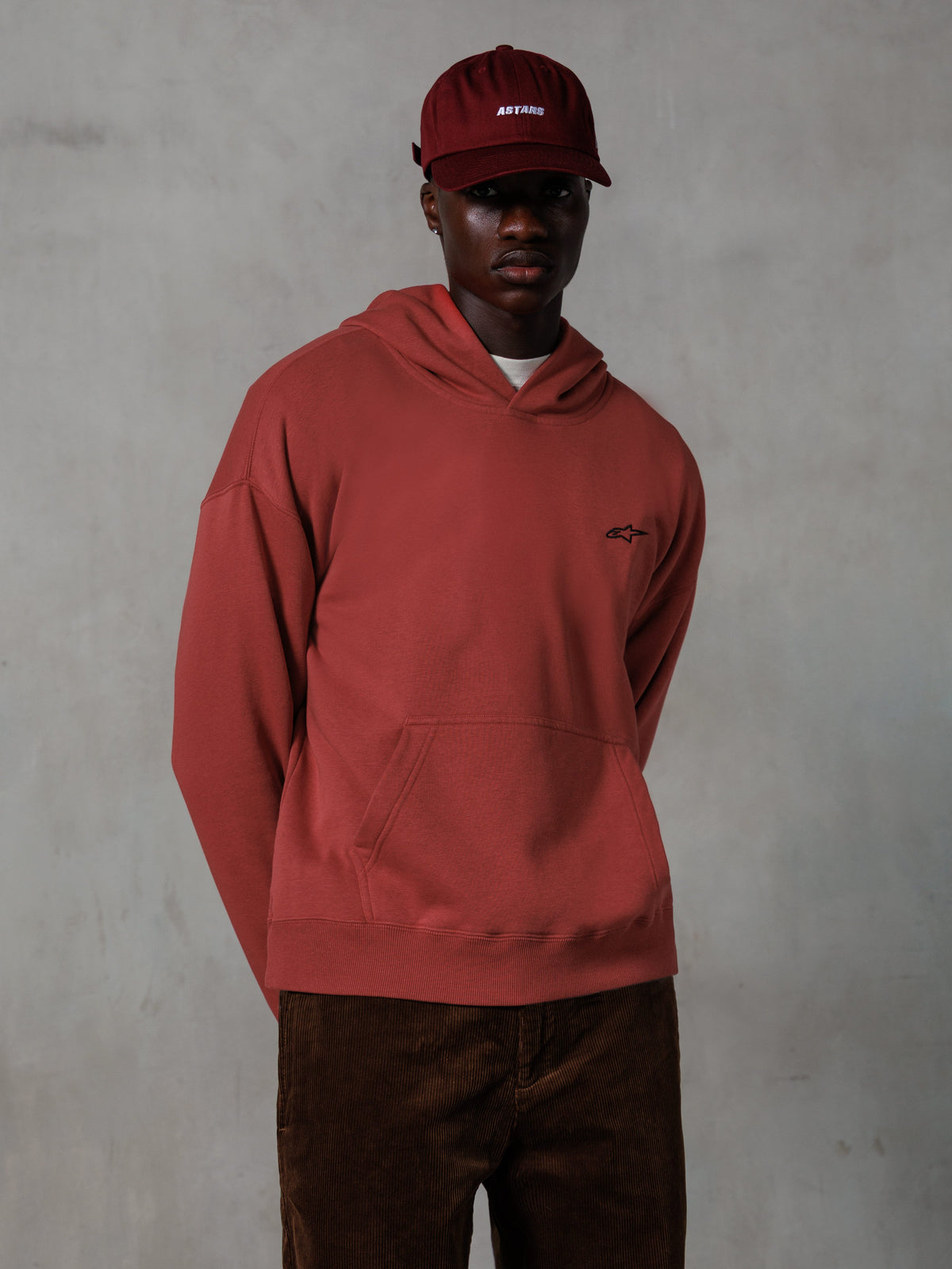 Alpinestars Essentials Ageless Hoodie, casual streetwear, baksteenrood, klein zwart logo op de borst, kangoeroezak aan de voorzijde, gedragen met een bijpassende rode ASTARS pet en bruine corduroy broek, strak minimalistisch ontwerp