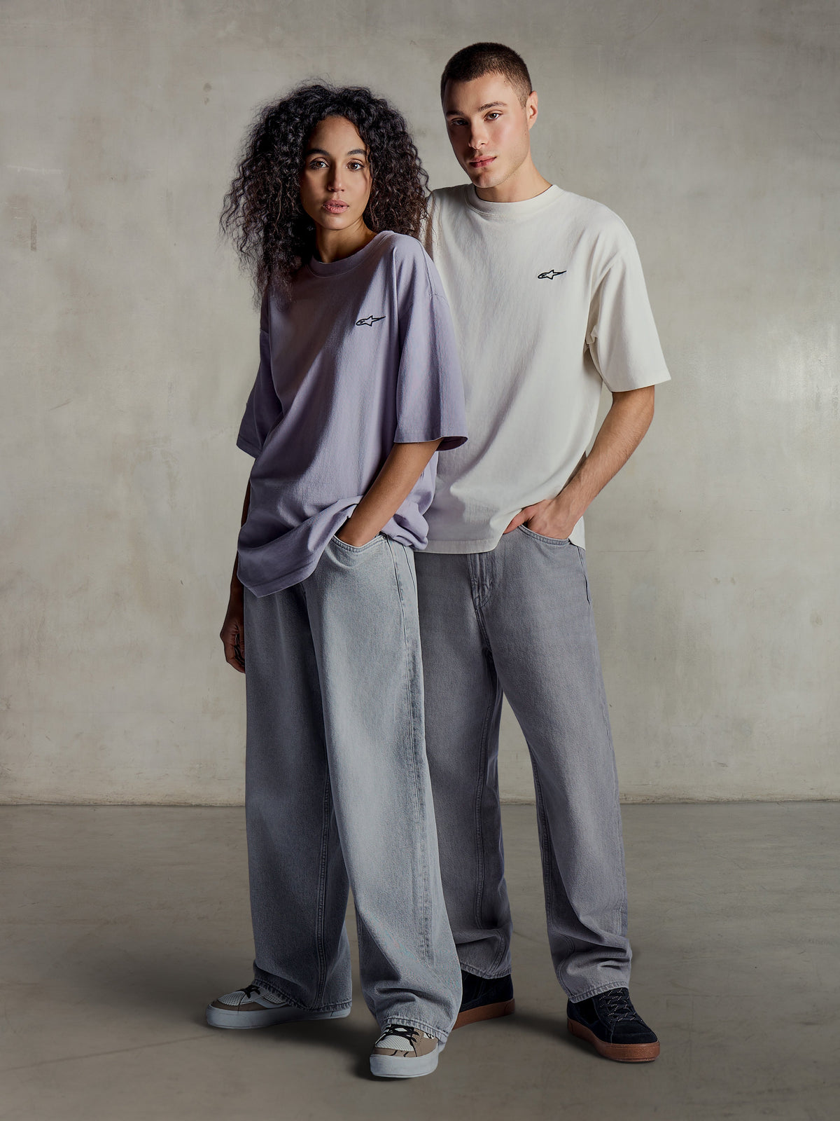 Alpinestars Essentials Ageless SS Tee, lifestyle-beeld van twee modellen in T-shirts met korte mouwen in lichtpaars en zilver, relaxed fit biologisch katoen met een subtiel logo op de borst, gecombineerd met wijde jeans voor een casual streetwear-stijl