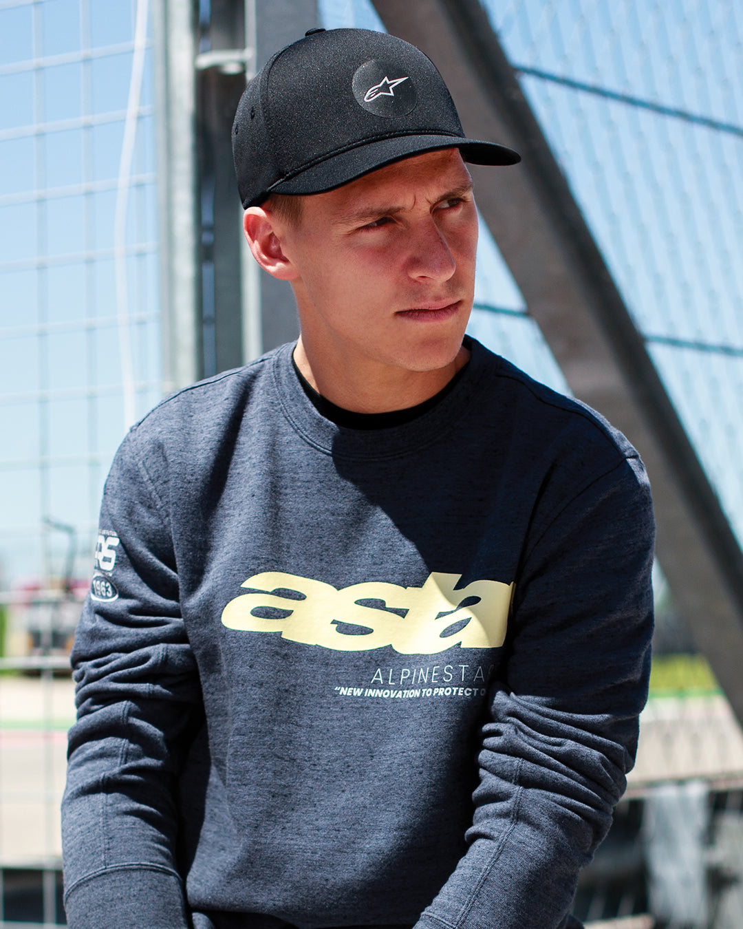 Fabio Quartararo draagt de Alpinestars Apex Pet in het zwart, een gestructureerde pet met gebogen klep en rond sterlogo, gecombineerd met een donkergrijs gemêleerd sweatshirt met gele branding, casual motorsport-geïnspireerde lifestyle.