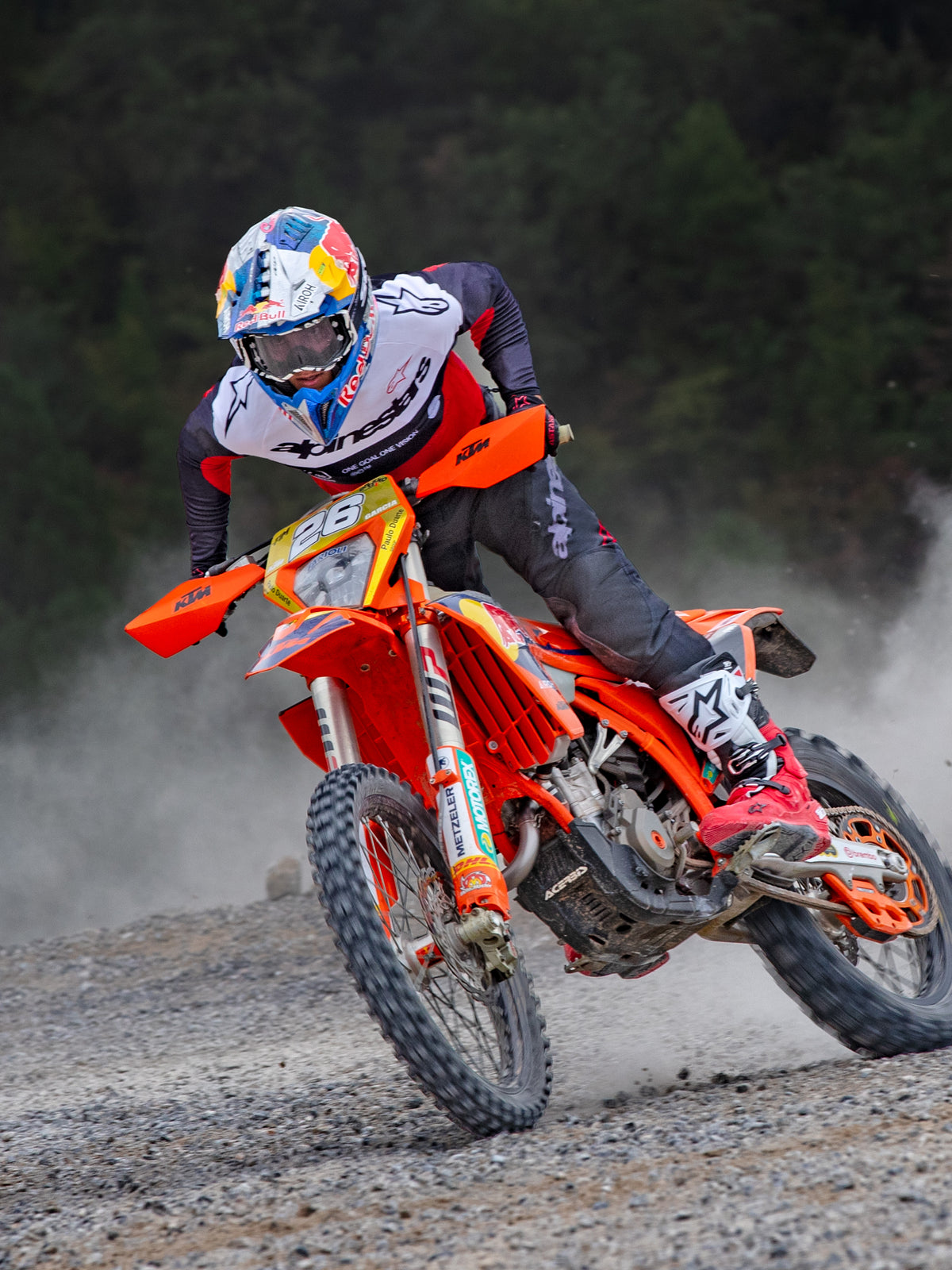 Motocrossrijder in actie op een oranje KTM-motor, gekleed in Alpinestars Pro-Dura-uitrusting met een rood, wit en zwart shirt, een donkergrijze broek, rood-witte laarzen en een Red Bull-racehelm, die een scherpe bocht maakt op een grindpad