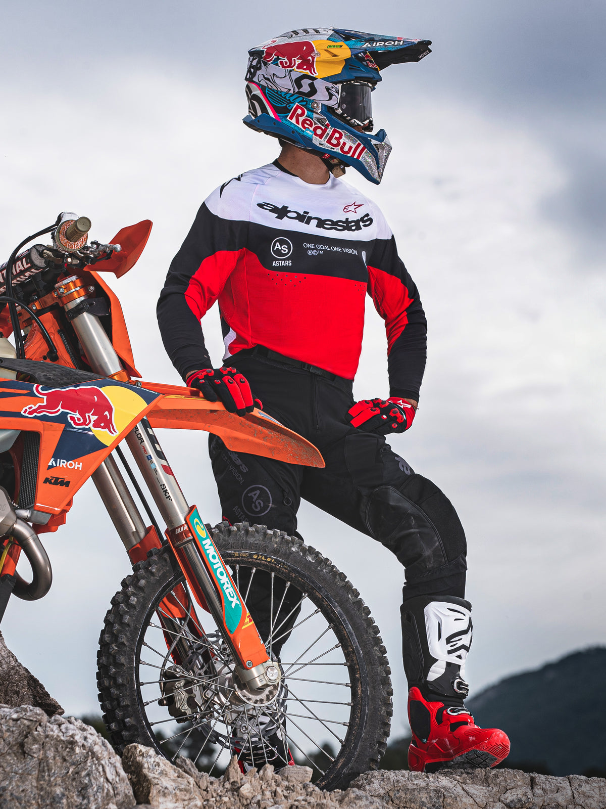 Motorcrosser staat naast een oranje off-road motorfiets op rotsachtig terrein, draagt Alpinestars Pro-Dura uitrusting bestaande uit een rood, wit en zwart jersey en een zwarte broek, gecombineerd met rood-zwarte handschoenen, wit-rode laarzen en een Red Bull-helm, complete professionele enduro-outfit.